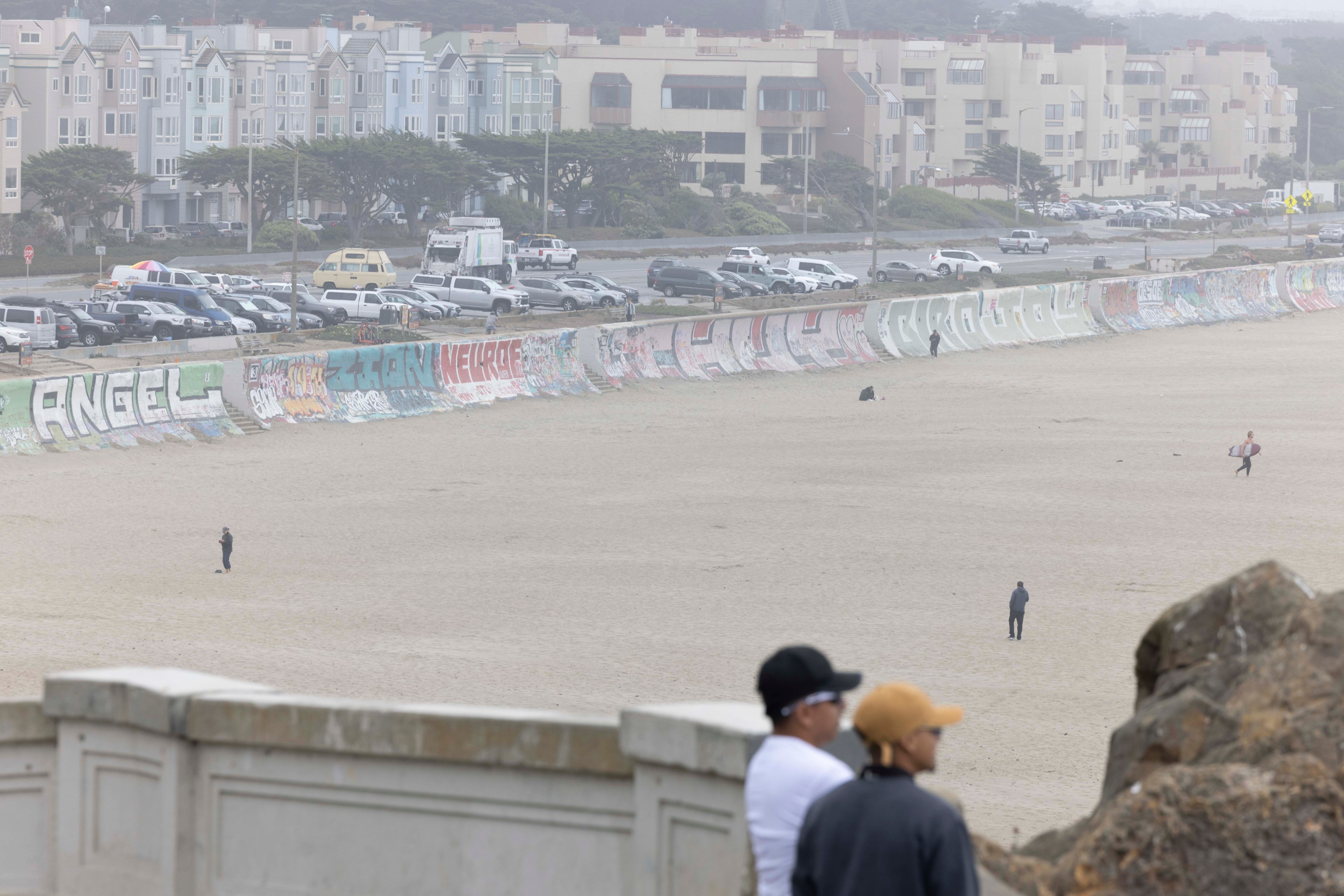 The image shows a sandy beach with a long wall covered in colorful graffiti. In the background, there are parked vehicles and multi-story residential buildings.