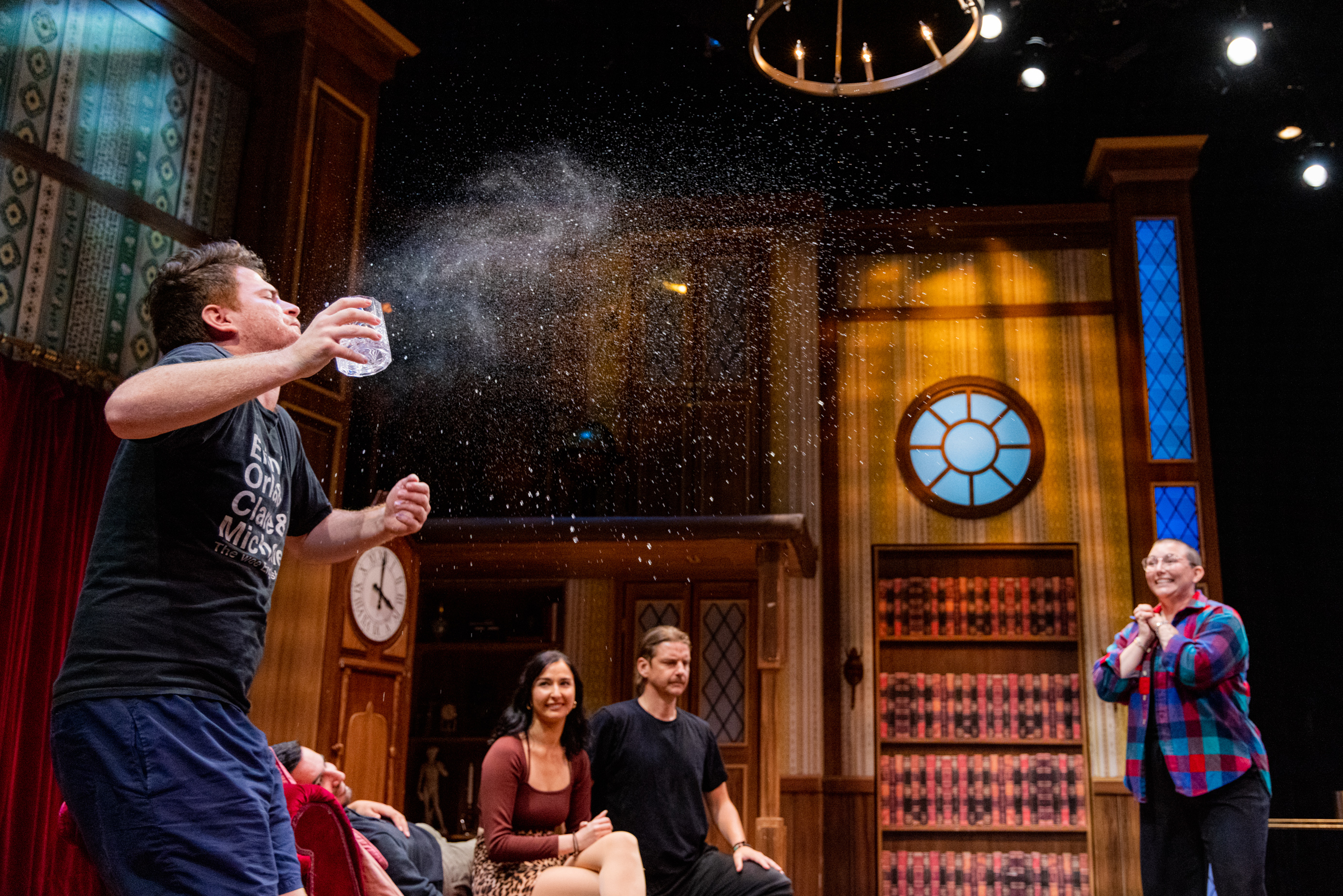 A man is spraying water from his mouth, as three other people, seated nearby, watch and laugh in a colorful, library-like setting with stage lights.