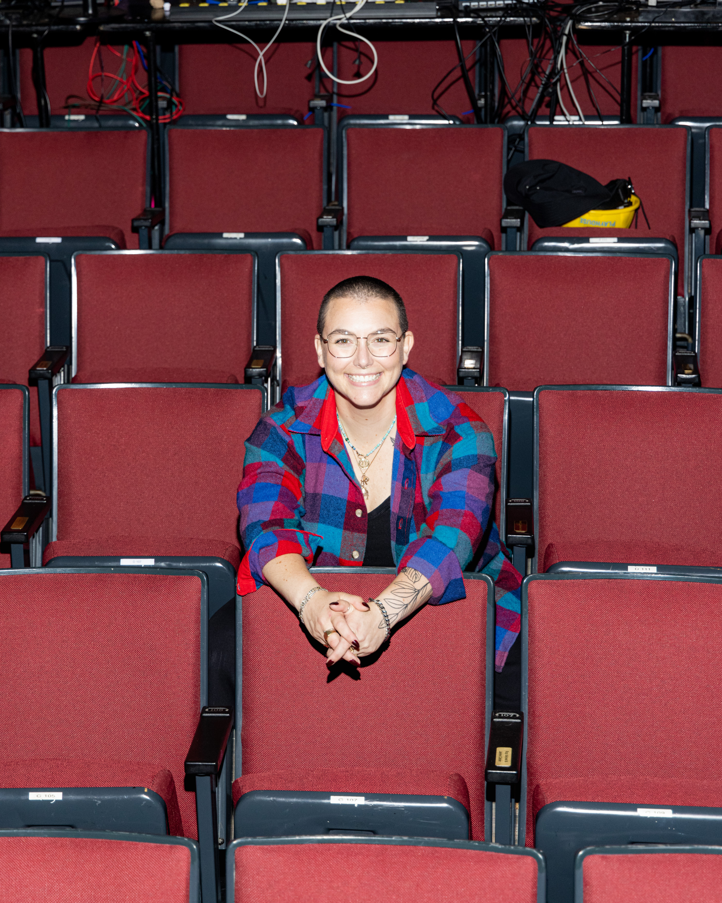 A smiling person with short hair and glasses, wearing a colorful plaid shirt, sits in an empty auditorium with red seats and cables overhead.