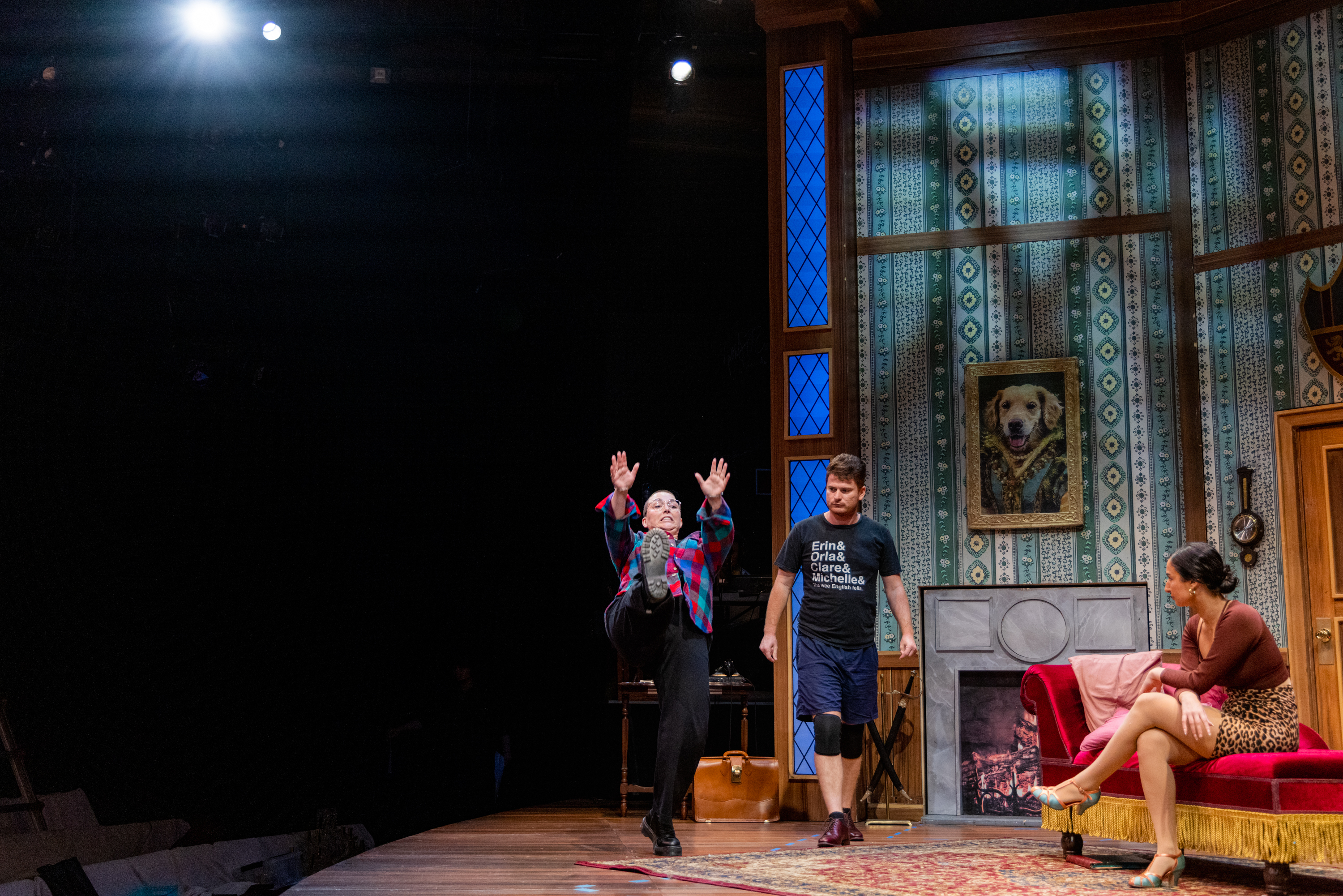 On a stage, one person in a plaid shirt energetically kicks a leg up, another in a black t-shirt and shorts stands nearby, while a woman in a leopard skirt sits on a red couch.