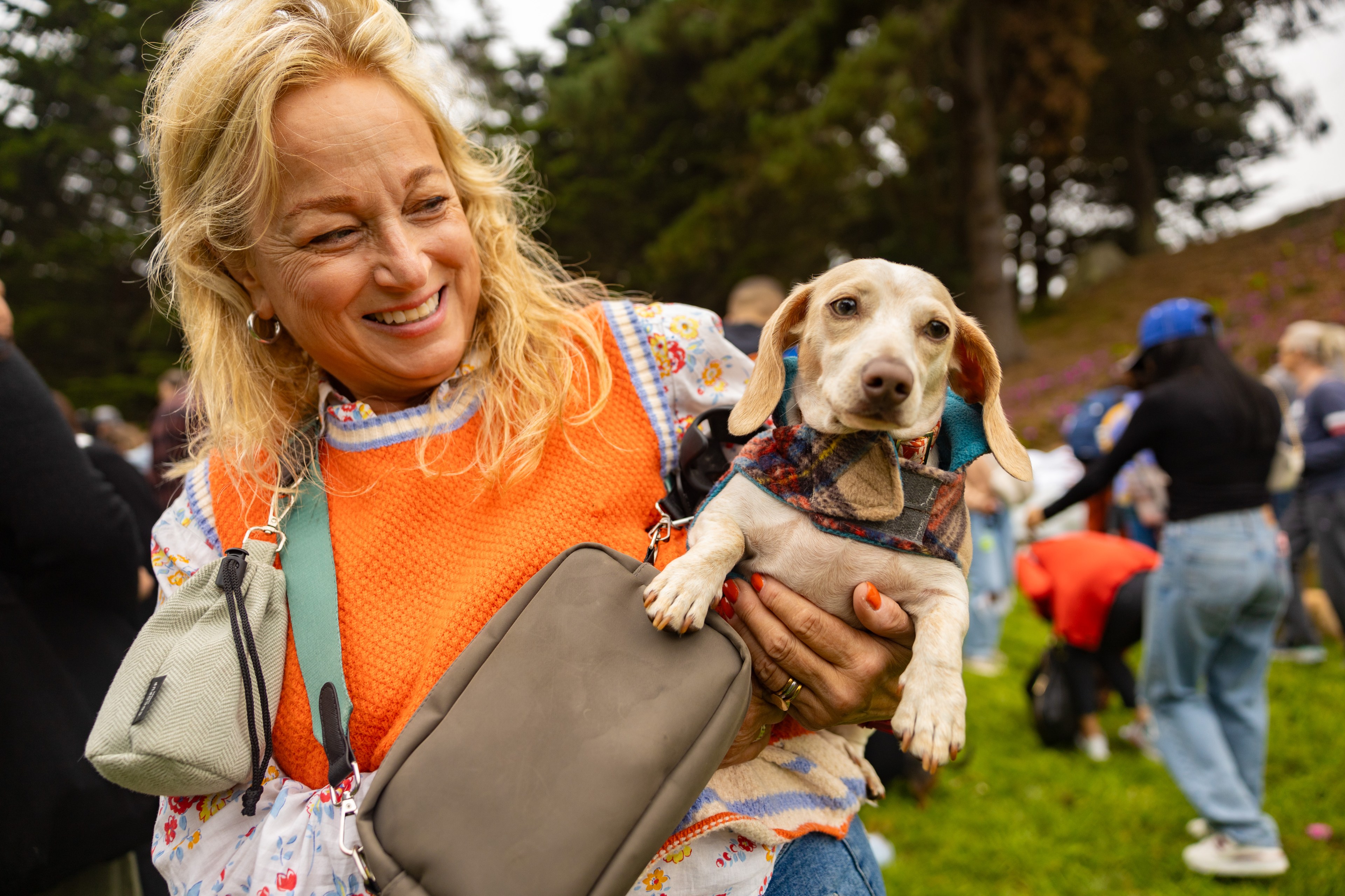 A smiling woman in an orange top holds a small dog dressed in a plaid outfit. They are outdoors with trees in the background and people around.