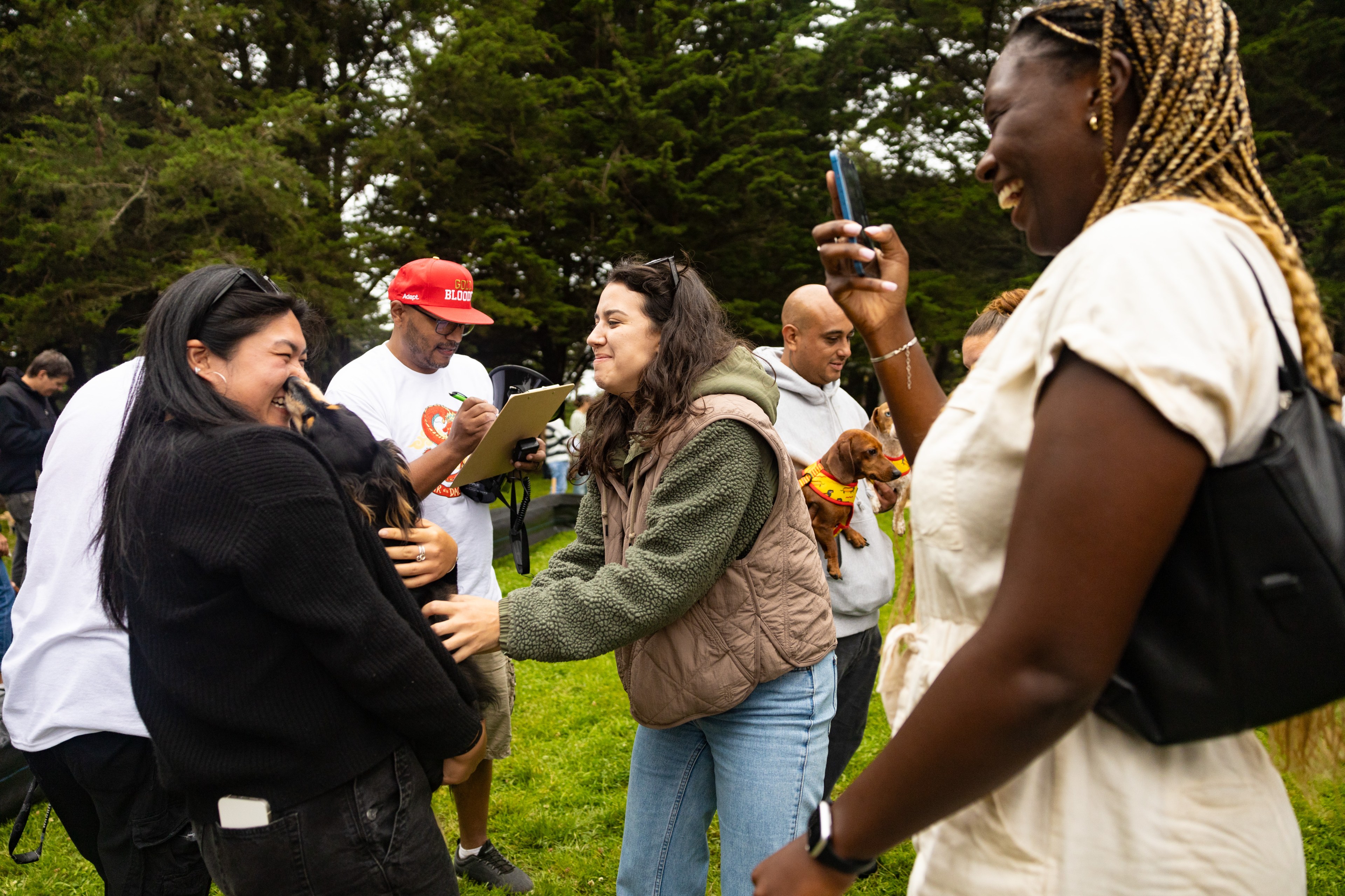 A group of people are outdoors in a park, smiling and interacting, with one woman holding a dog. Another person is taking a photo while others watch and laugh.