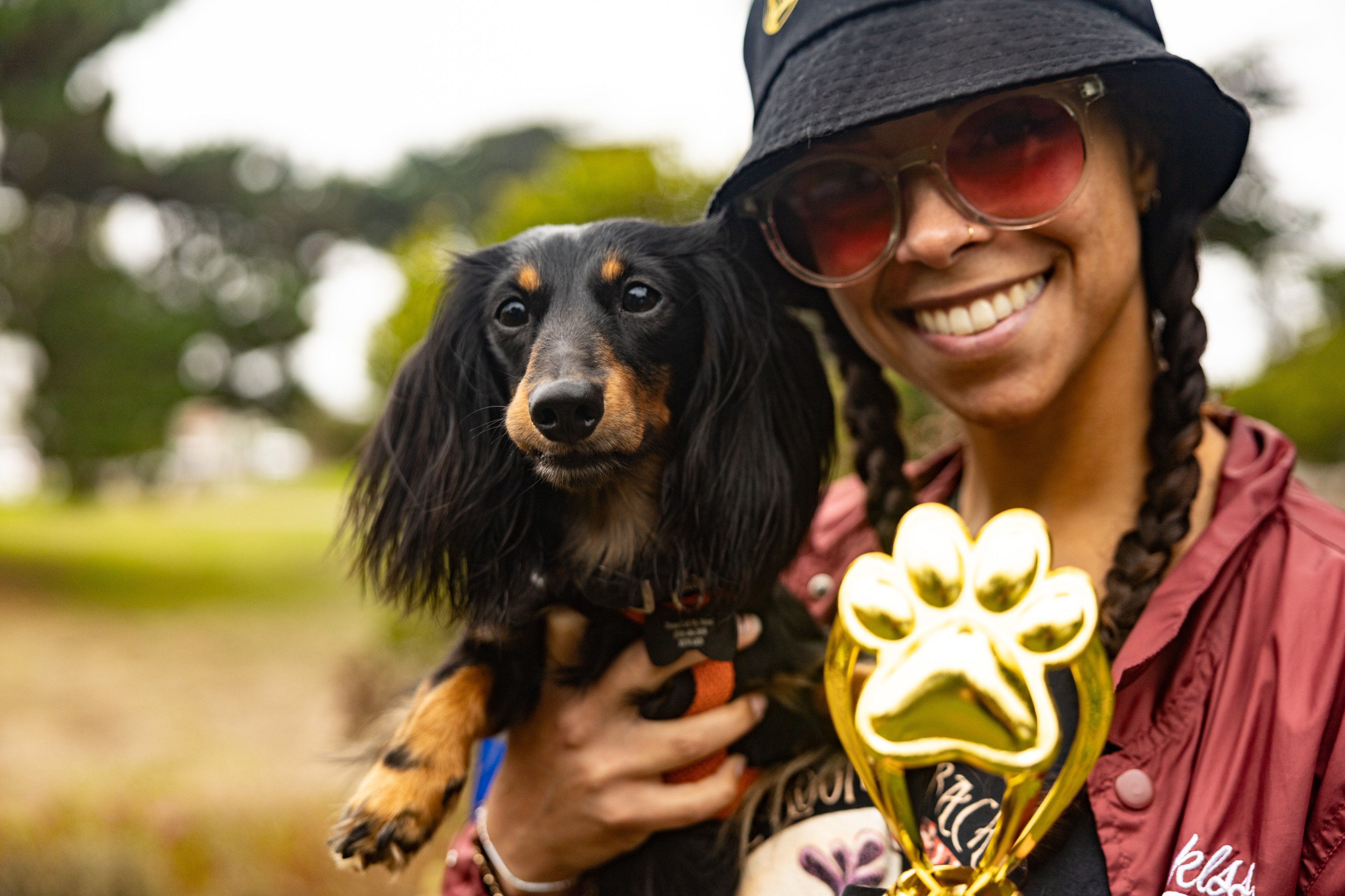 A smiling person wearing sunglasses and a hat holds a long-haired black and tan Dachshund, and a gold paw-shaped trophy, with a park background visible.