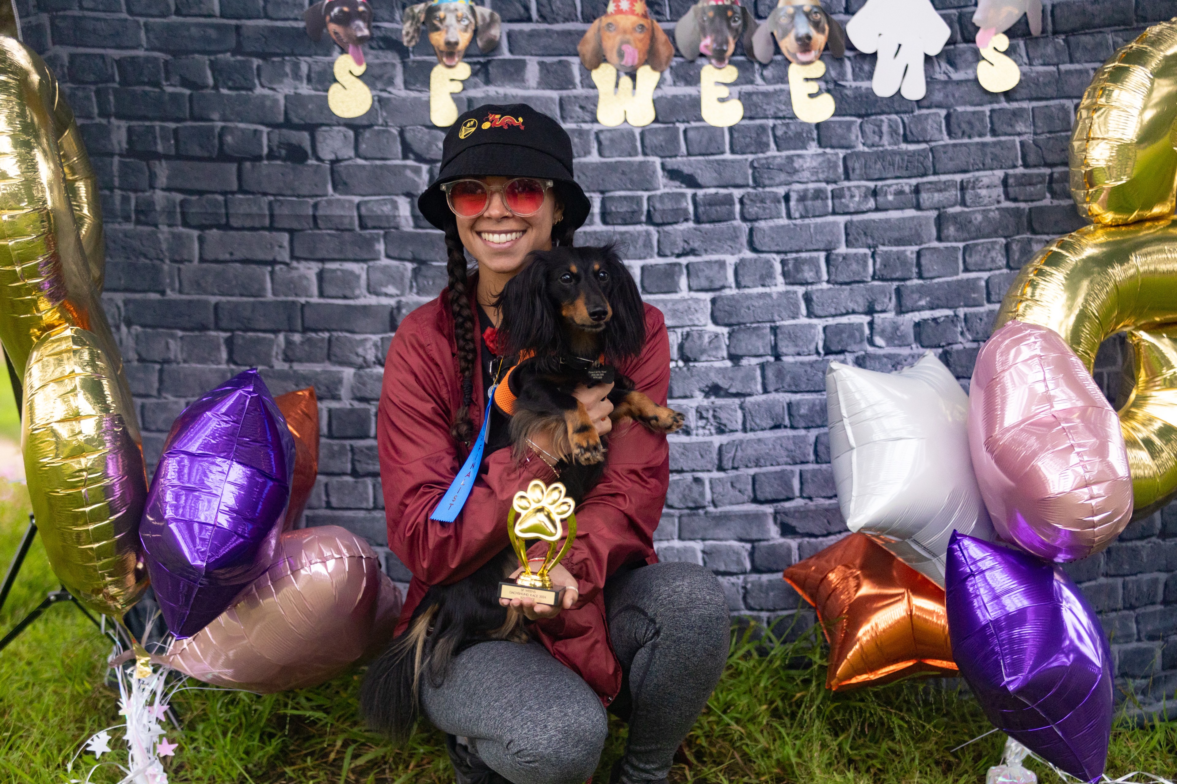 A smiling person in a red jacket and black hat holds a black dog and a gold paw-shaped trophy, surrounded by colorful balloons and a &quot;SFWEE&quot; backdrop with dog photos.