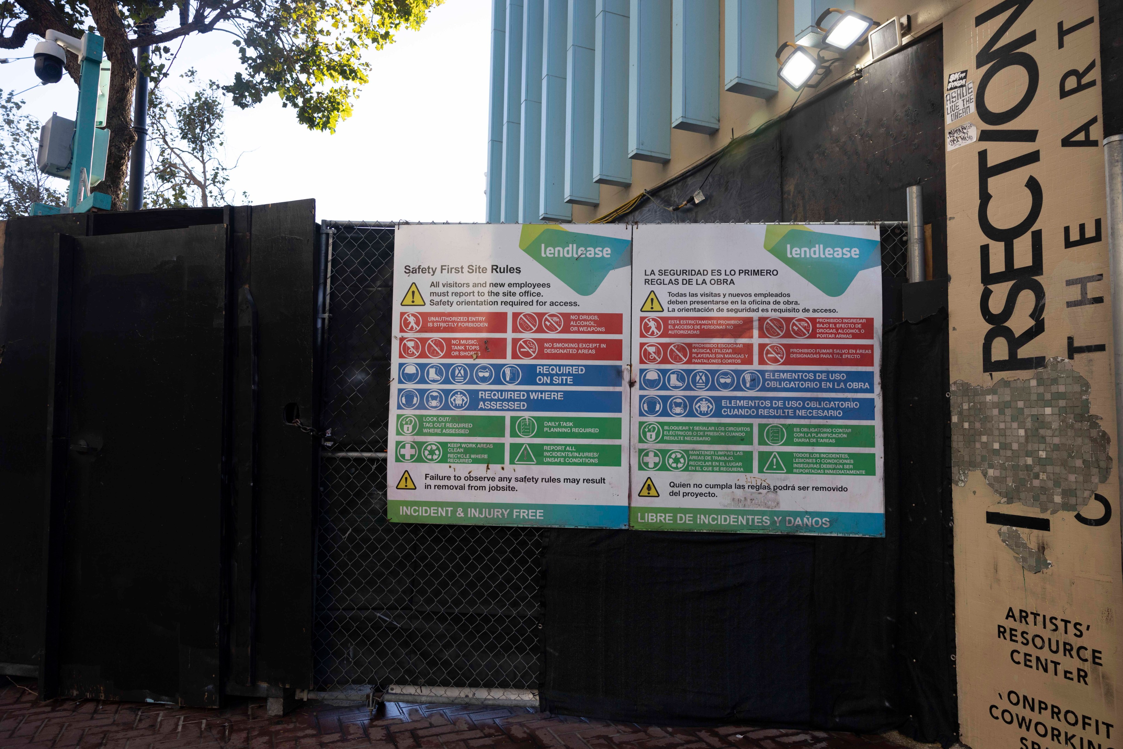 The image shows a construction site entrance with black gates, surveillance cameras, and safety signs in English and Spanish, indicating site rules and required safety measures.