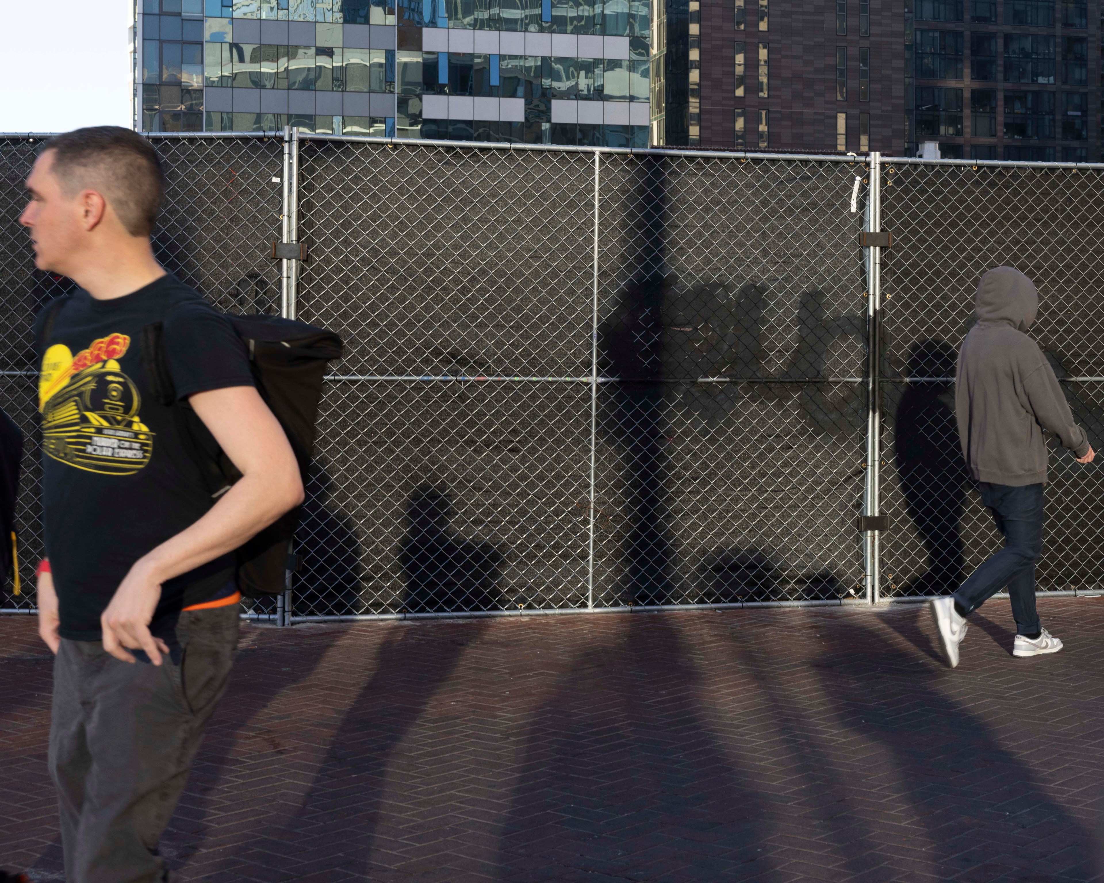 Two people are walking in front of a tall chain-link fence with black privacy fabric; one wears a dark T-shirt and backpack, the other is in a gray hoodie, jeans, and sneakers.