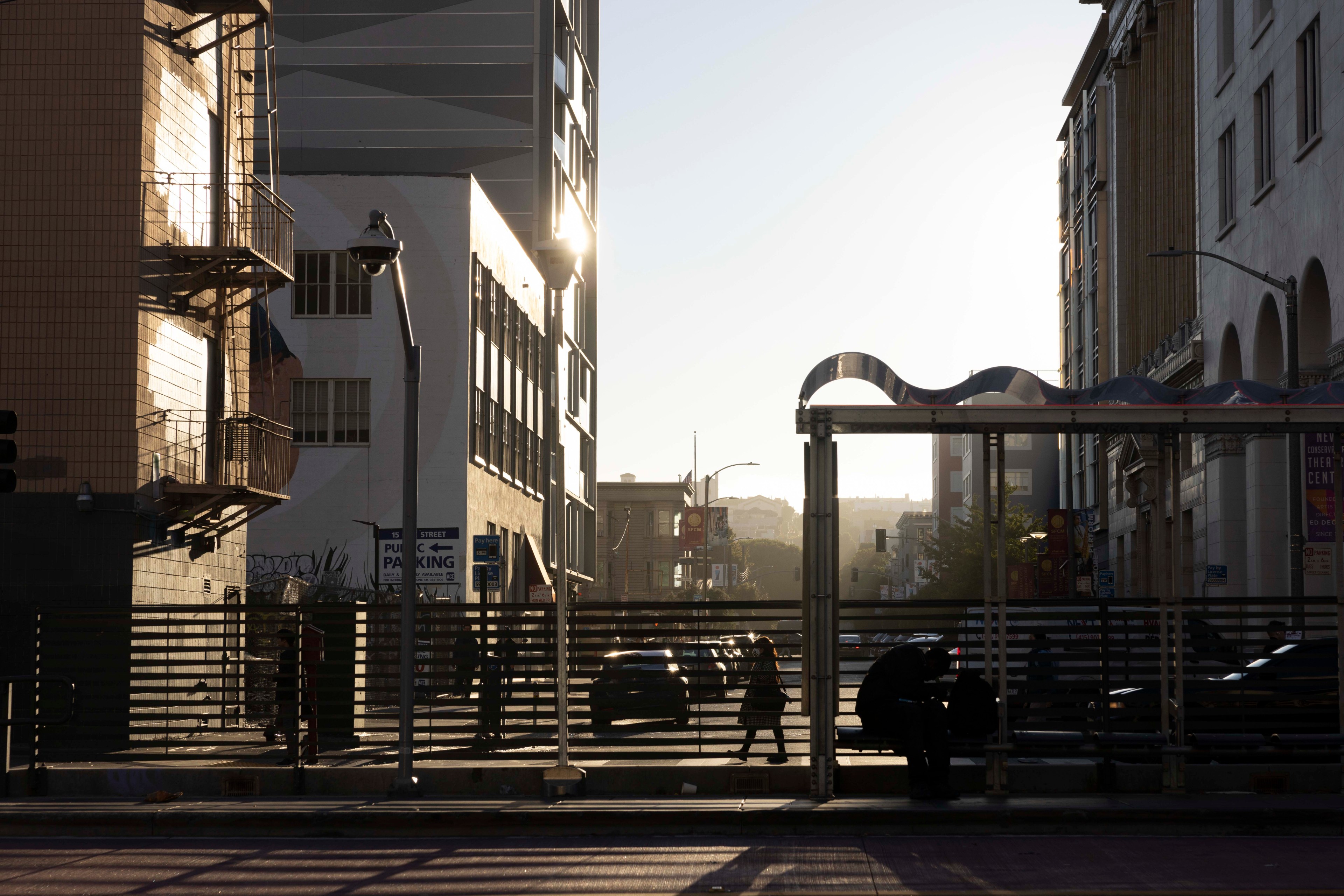 The image shows a city street at dawn or dusk with a golden light casting shadows. There are buildings, a streetlight, people walking, and a bus stop in silhouette.