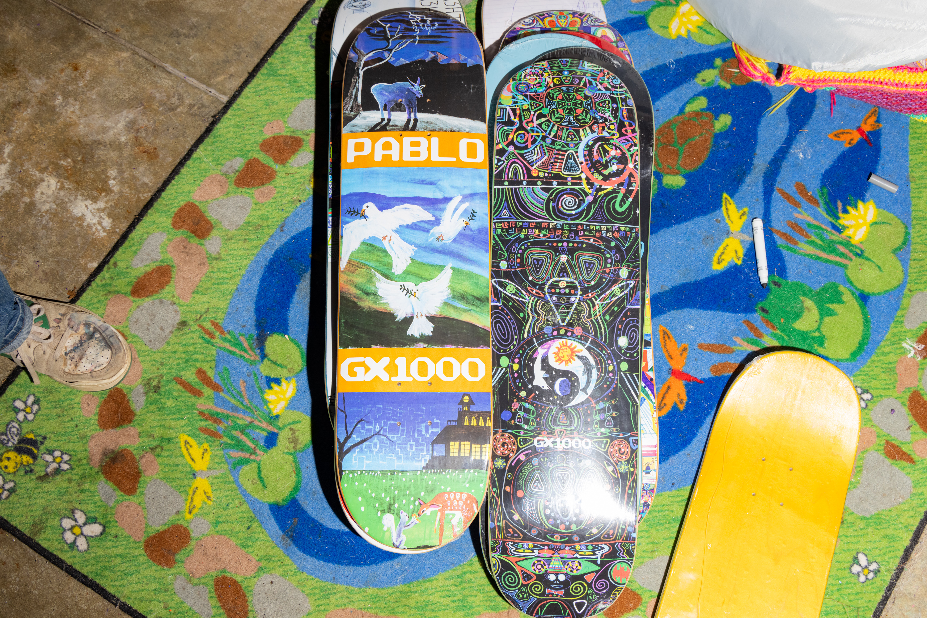 The image shows two skateboards with colorful designs, the left one features animals and &quot;PABLO GX1000,&quot; while the right one has intricate patterns. They rest on a vibrant rug.