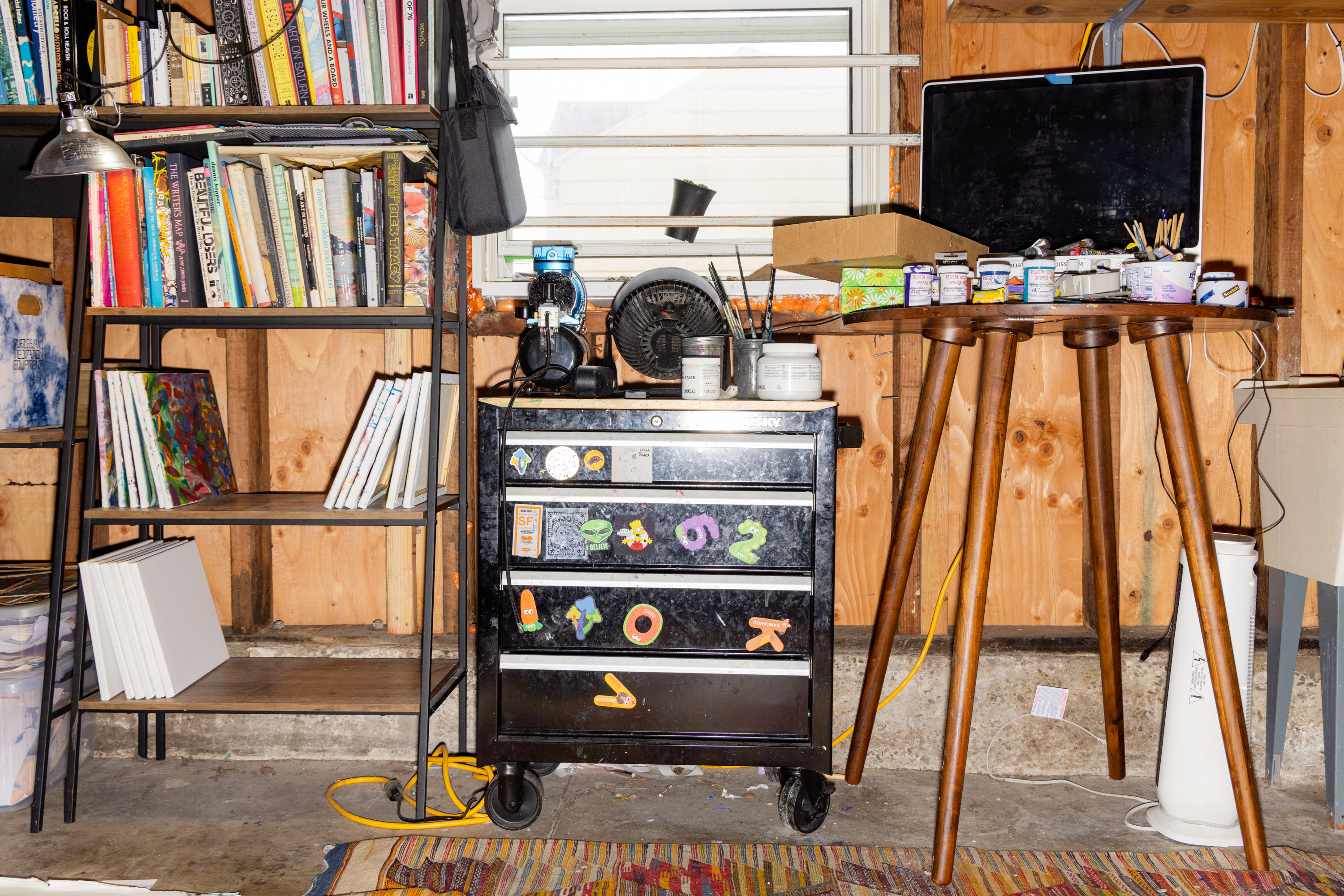 The wooden-walled room includes a bookshelf, art supplies, and a tool cabinet. It also contains a small table cluttered with paint cans and brushes, under a window.