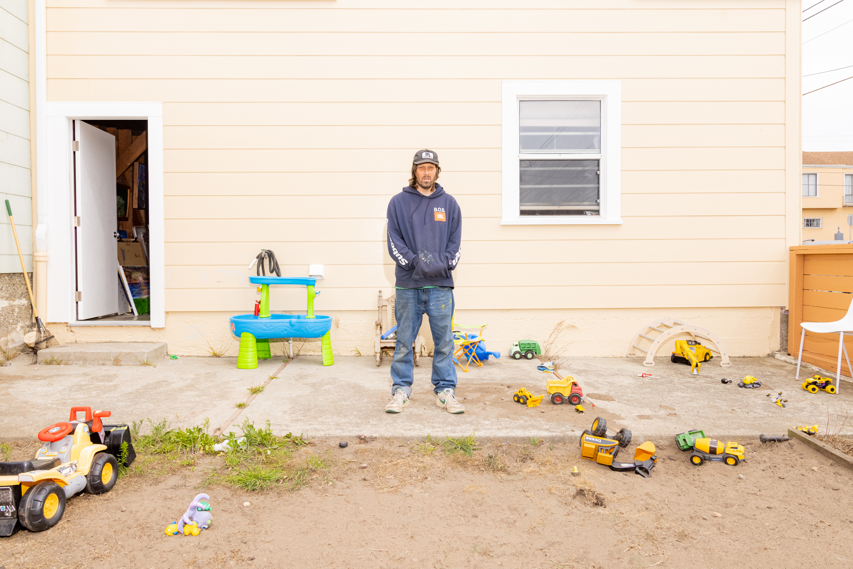 A person stands in front of a beige house with scattered children's toys and a blue table in the yard. There's a window and an open door with more items visible inside.