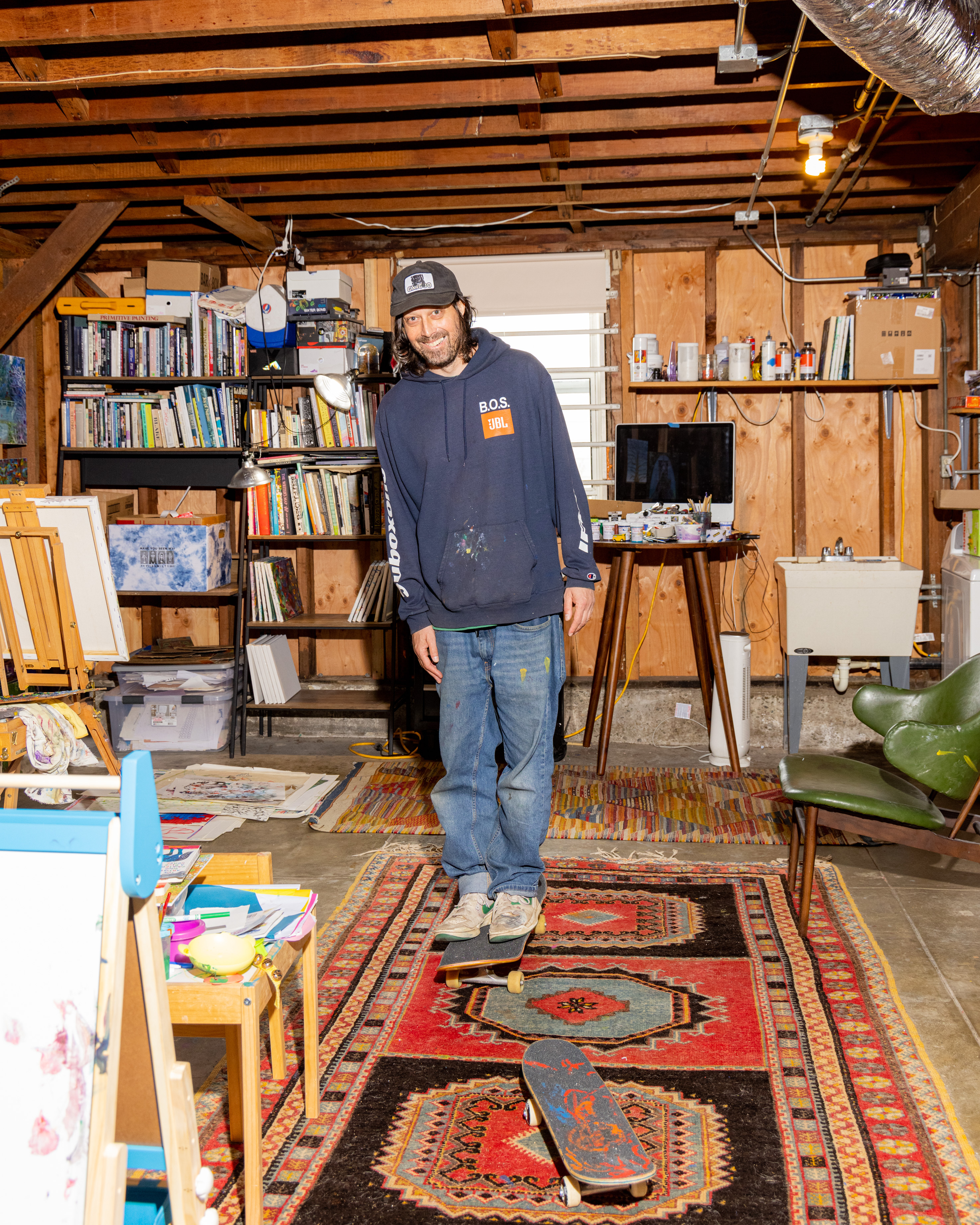 A man stands on a skateboard inside a cluttered room with bookshelves, rugs, art supplies, and a computer, smiling with his hands in his hoodie pockets.