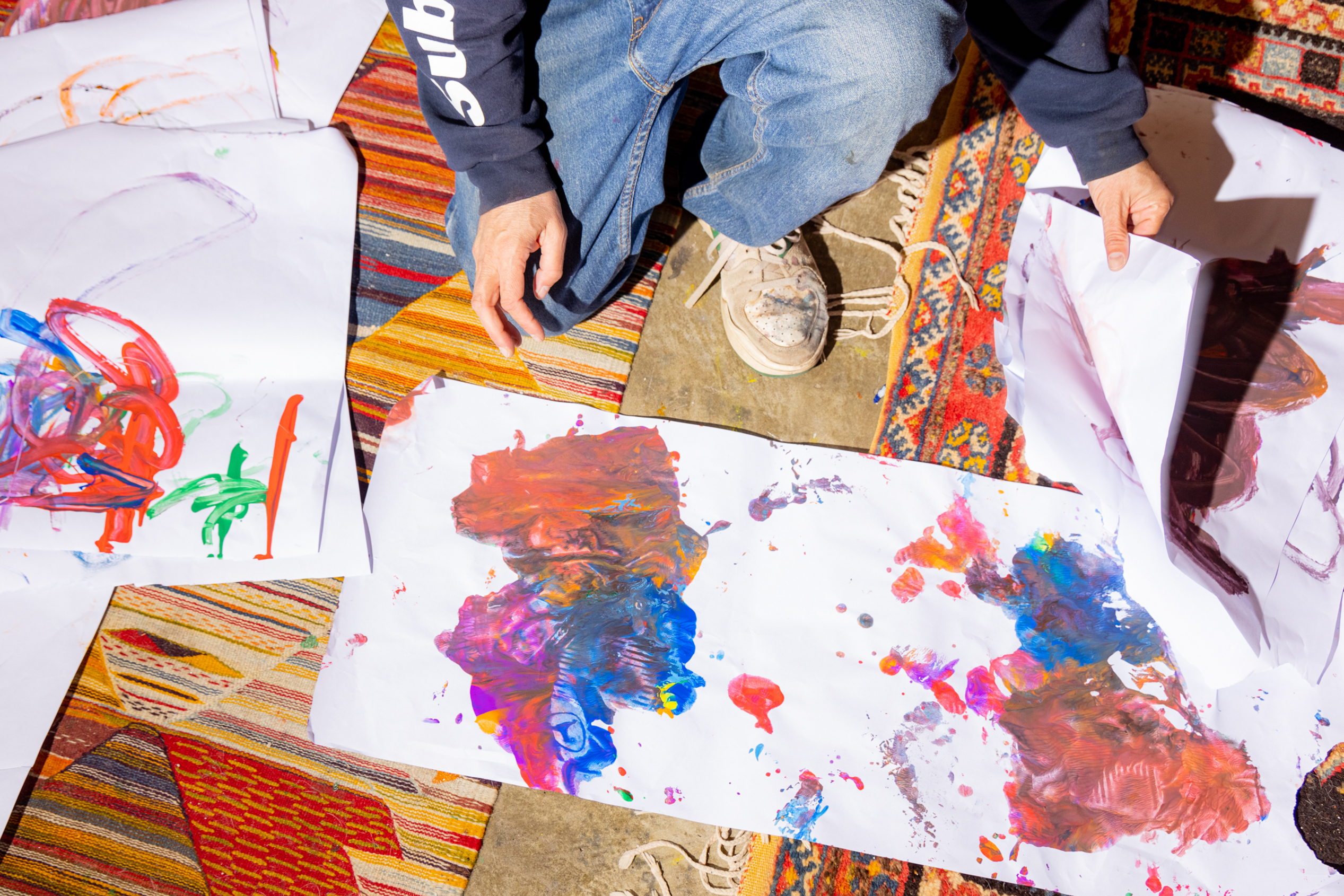 A person in jeans and a dark sweater sits on a colorful rug, surrounded by abstract, vibrant paintings on paper scattered on the floor.