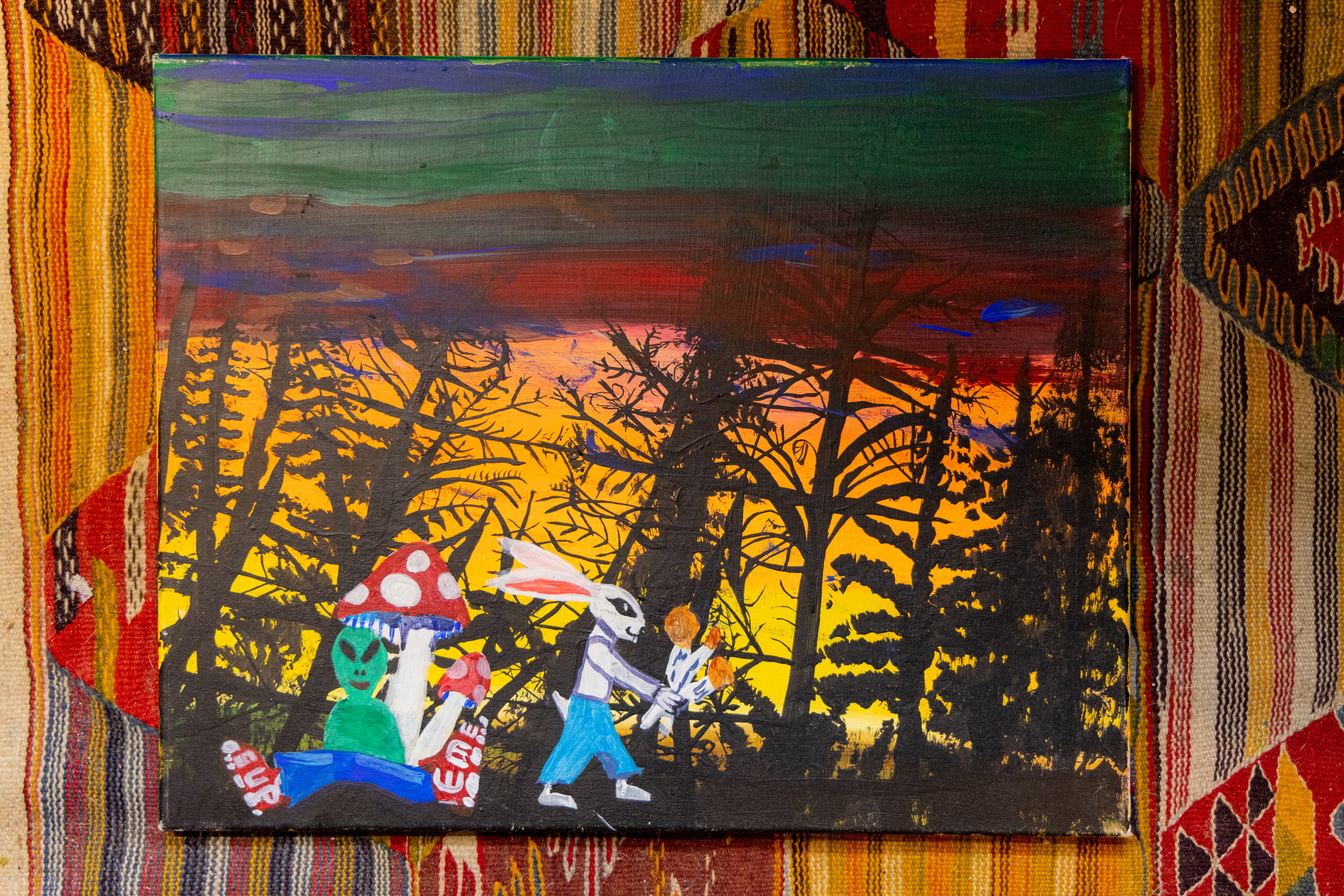 A colorful painting depicts a surreal scene of an alien and a bunny carrying a pumpkin, set against a backdrop of a sunset with silhouetted trees and mushrooms.