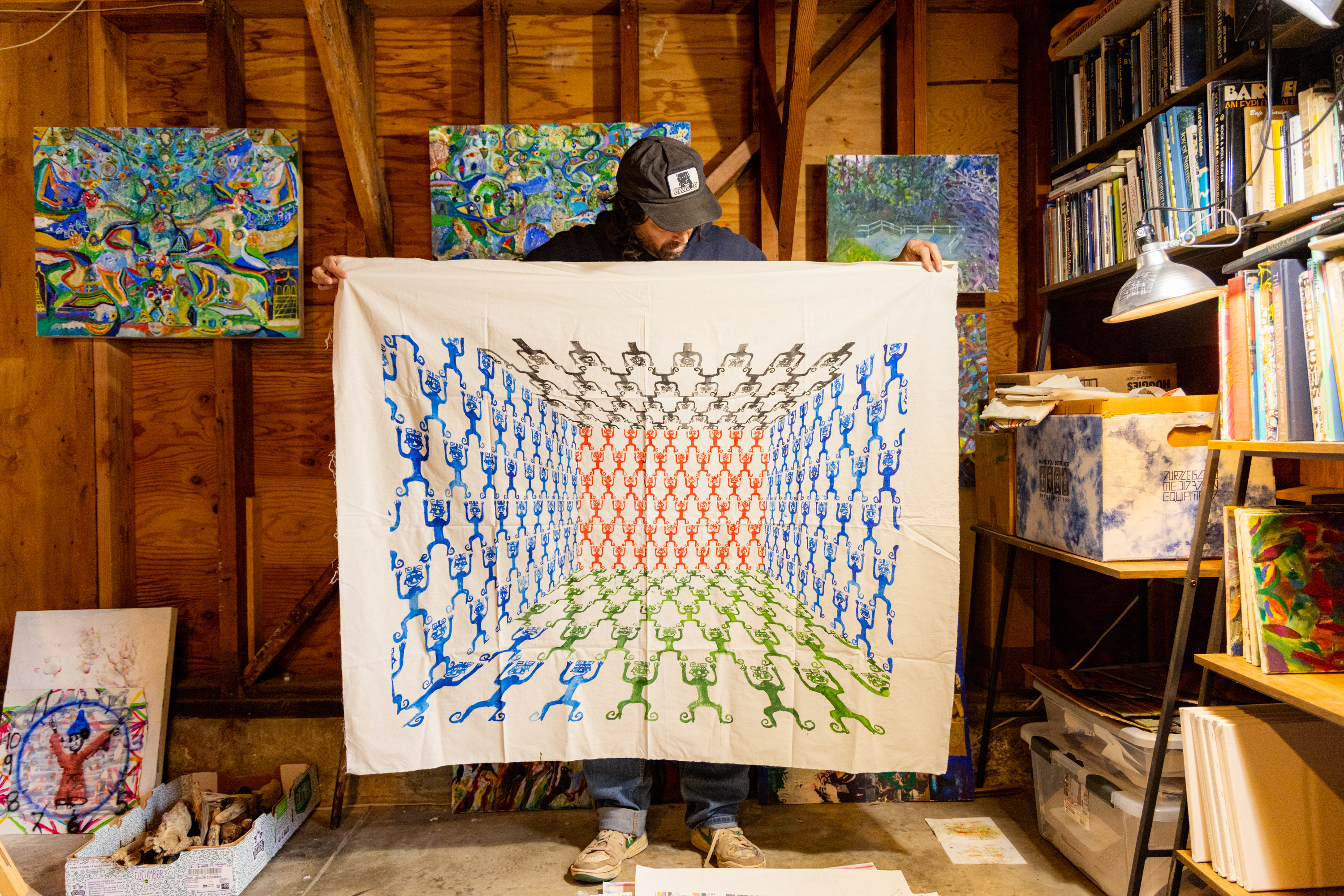 A person in a cap holds up a large, colorful fabric with intricate symmetrical patterns in blue, red, green, and black, standing in an art-filled studio or workshop.