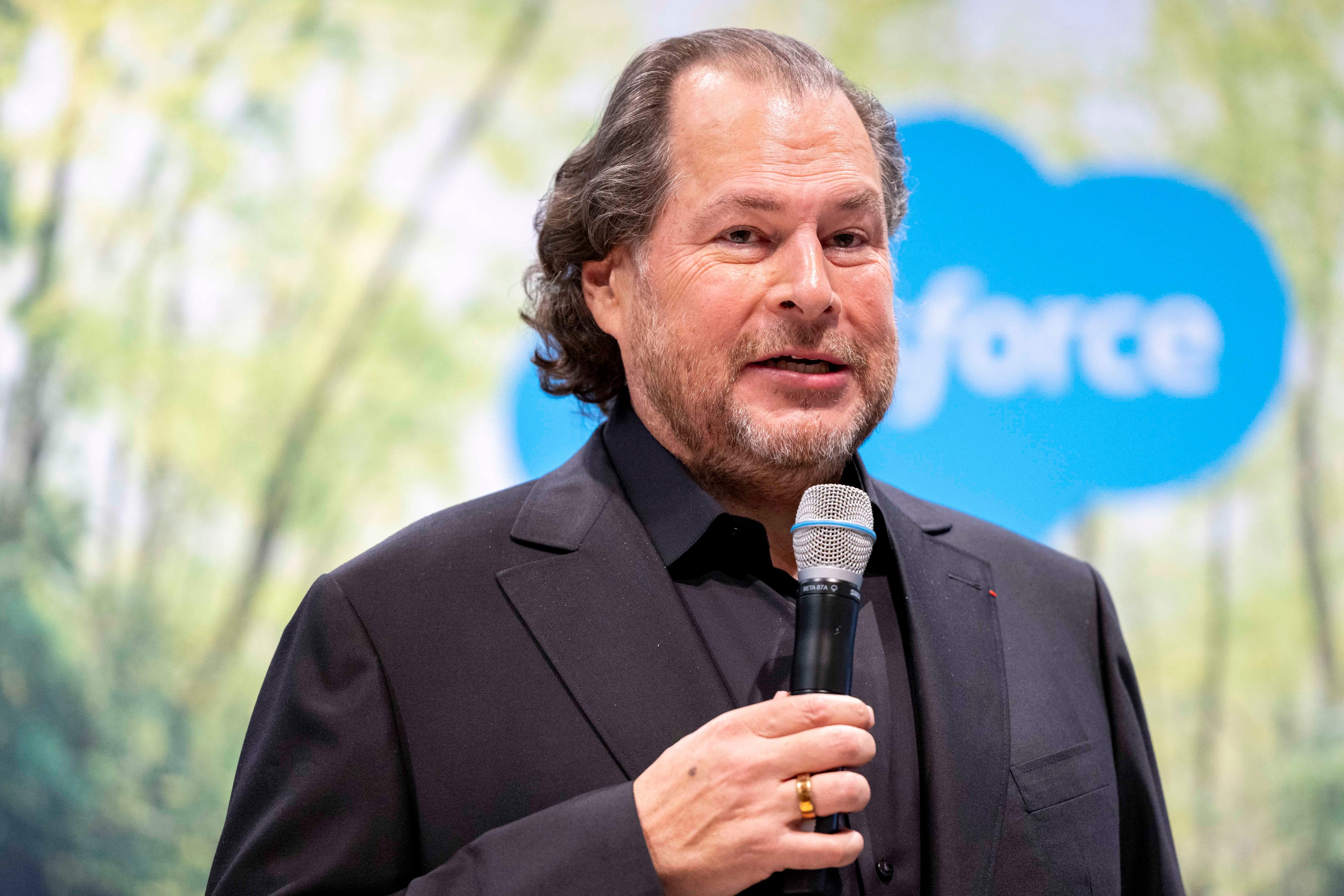 A man in a black suit holds a microphone, speaking against a blurred background with a blue logo partially visible. He has short, wavy hair and is smiling slightly.