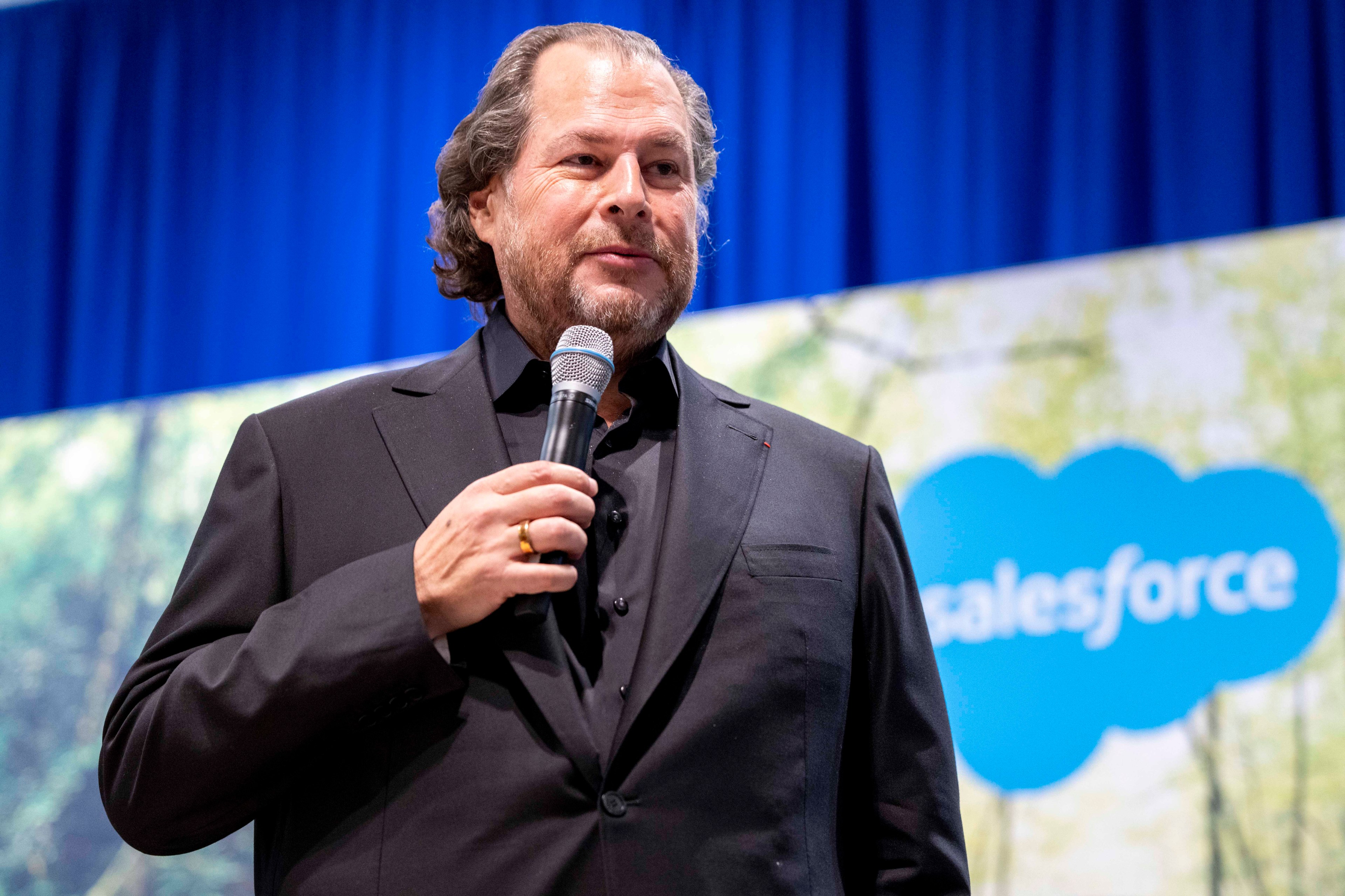 A man in a black suit is speaking into a microphone on stage. Behind him is a blurred blue and green background featuring a cloud-shaped Salesforce logo.