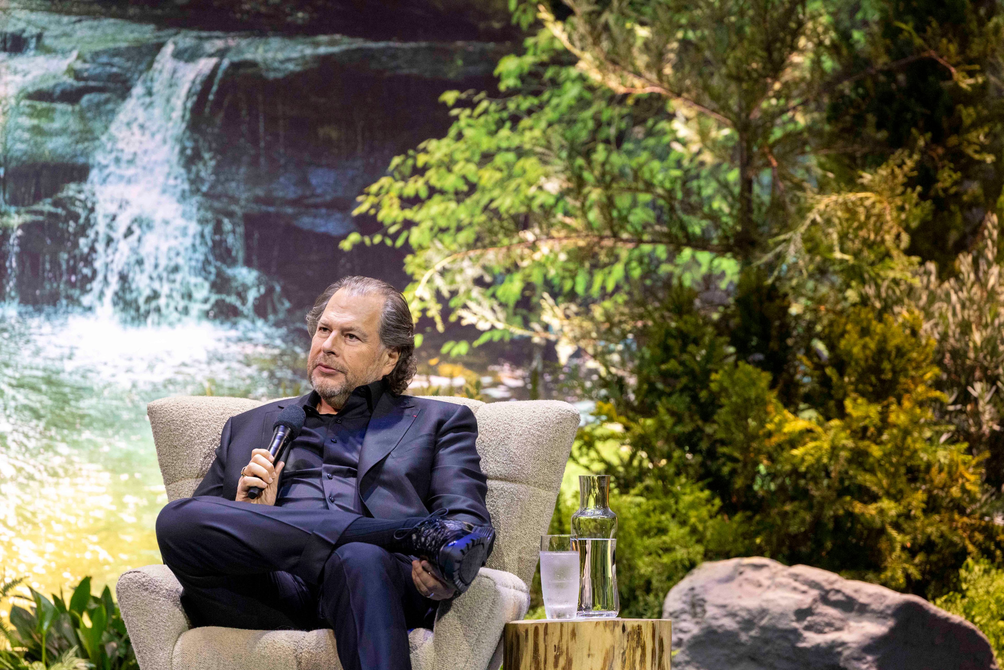 A man in a black suit sits in a chair, holding a microphone, with a lush, green forest and a waterfall backdrop behind him. A table with water and a glass is beside him.