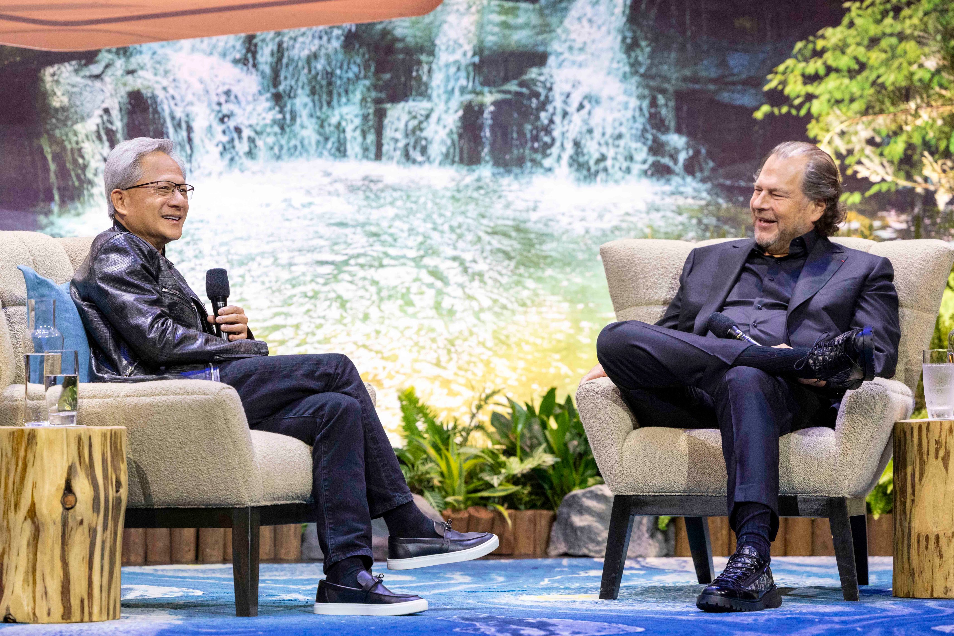 Two men sit and talk in armchairs with microphones, against a waterfall backdrop, next to small wooden side tables, with plants around. Both are smiling warmly.