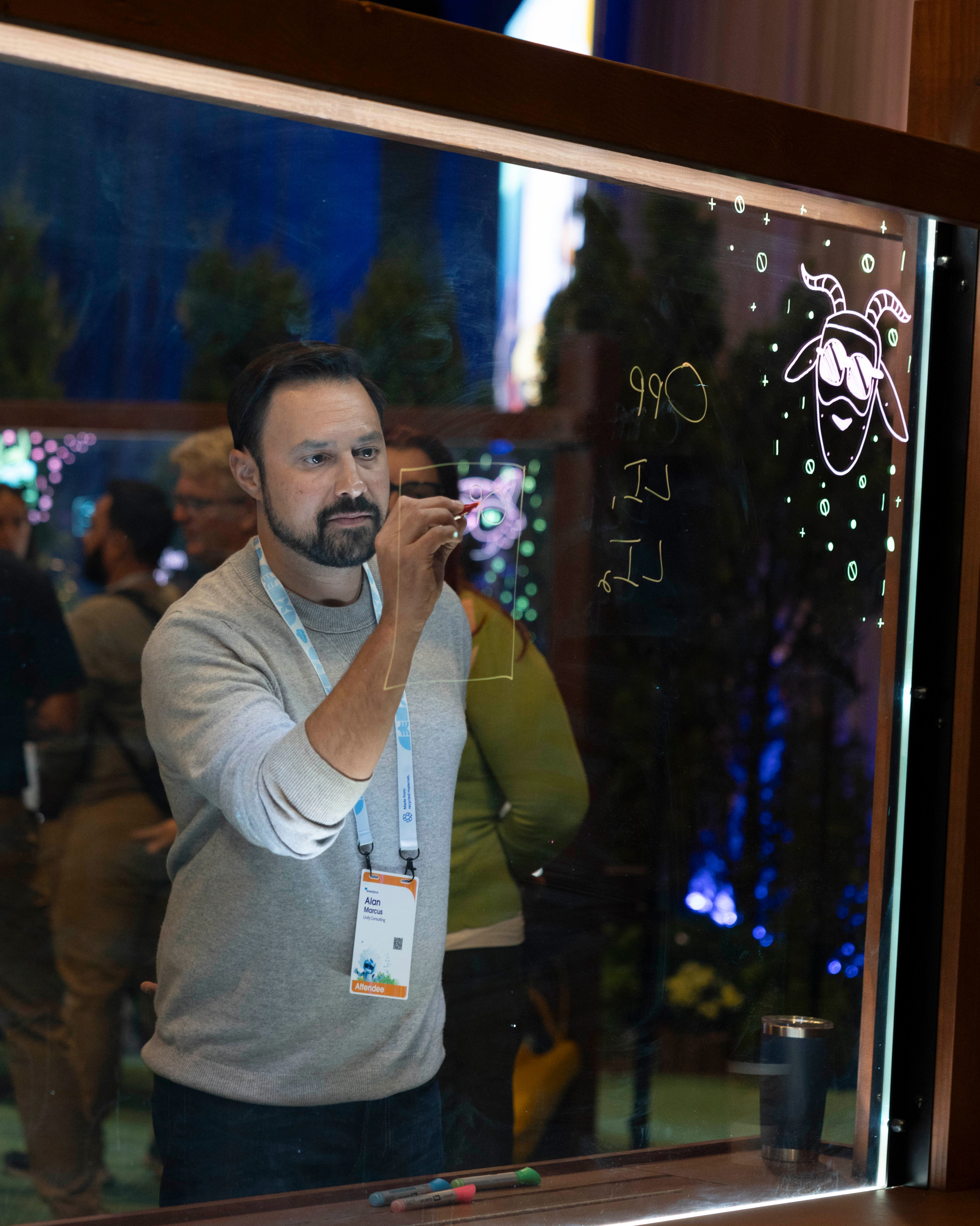 A man with a beard writes on a transparent board using markers. He wears a badge and is surrounded by an indoor, possibly conference, setting with other people and lights.