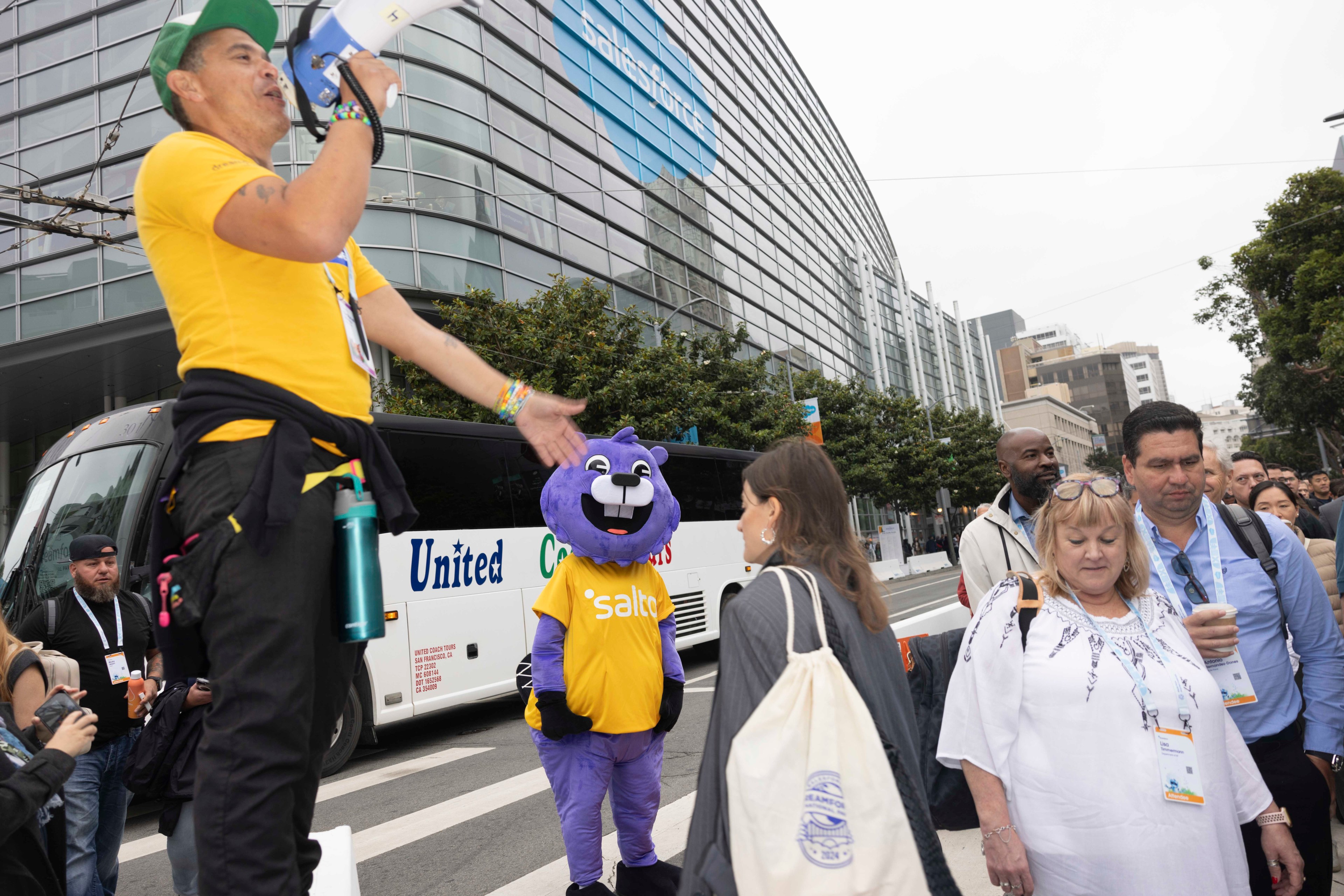 A man in a yellow shirt and green cap speaks into a megaphone, while a person in a purple mascot costume stands nearby. People walk past a bus in front of a large building.