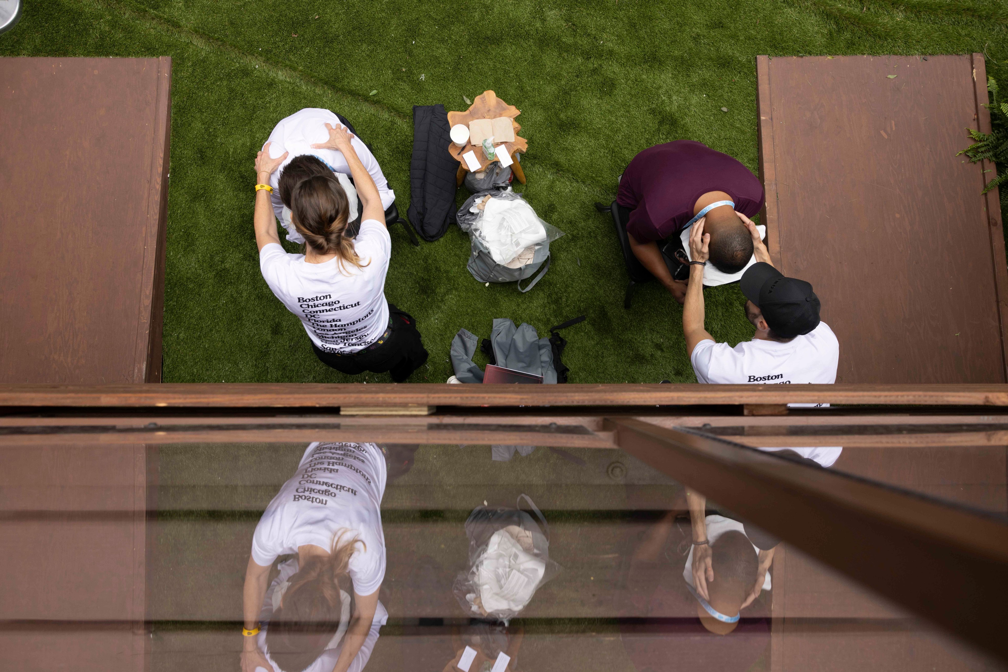 Two people are receiving massages on a grassy area, with two others standing behind them. A small wooden table with various items is placed between them.