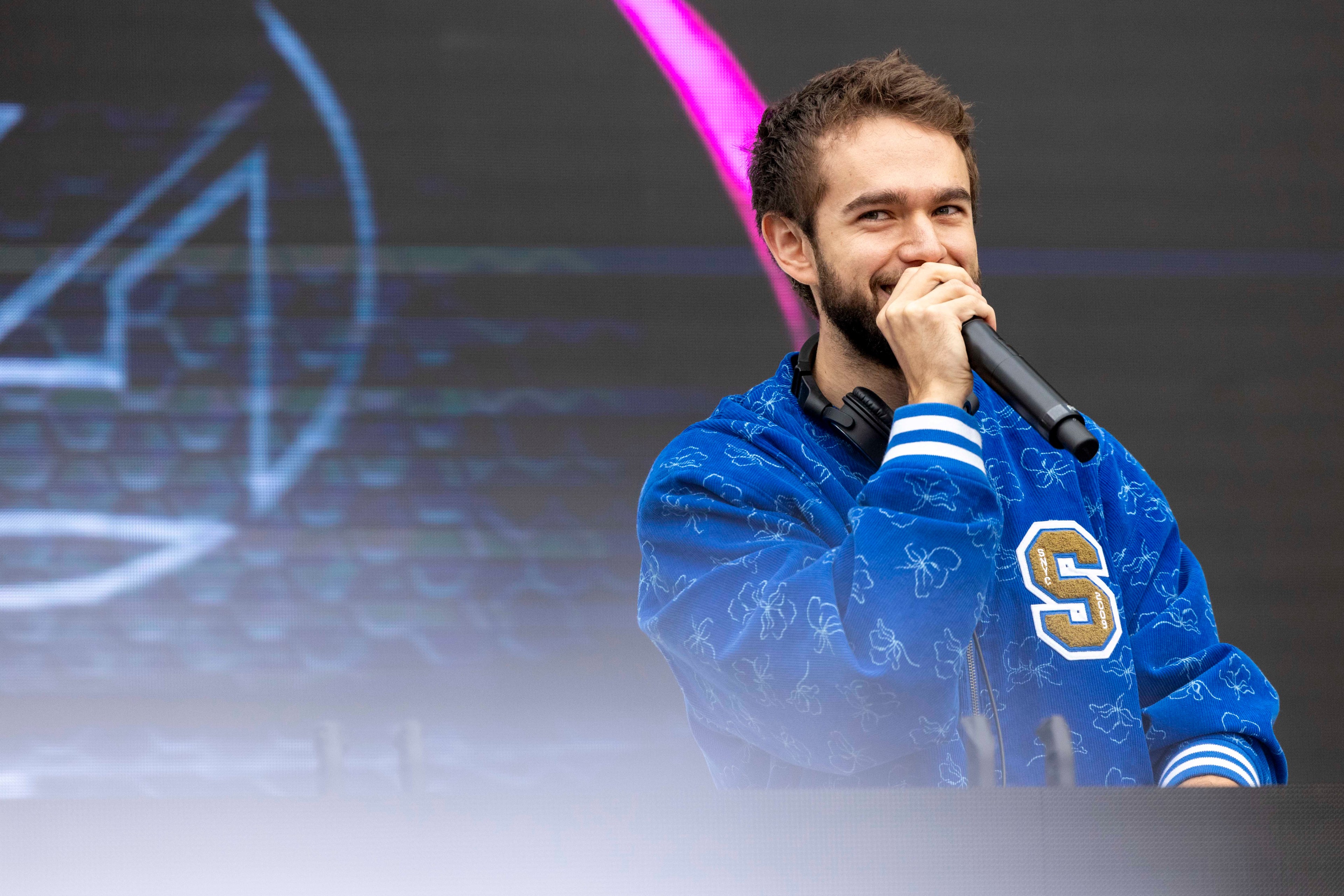 A man in a blue jacket with a large &quot;S&quot; emblem, holding a microphone, smiles on stage. A dark background with faint blue and pink lights surrounds him.