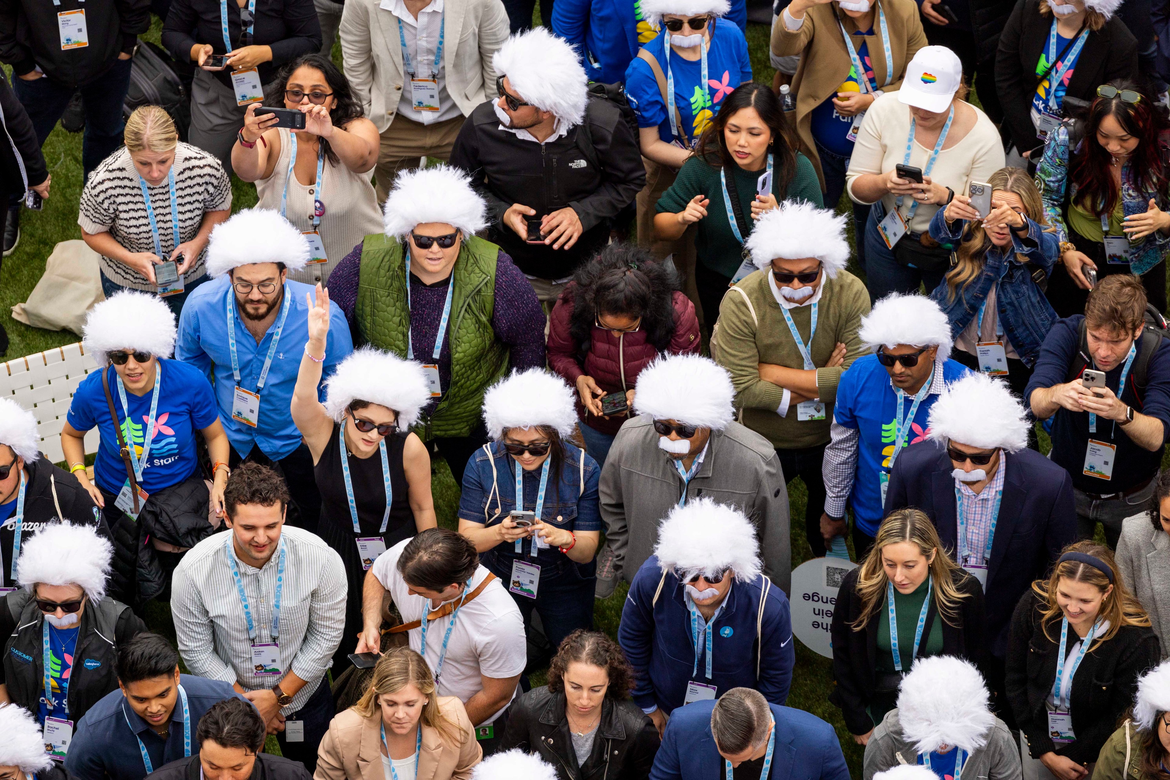 A group of people outdoors, many wearing white wigs and fake mustaches, are looking at their phones or cameras. Some are raising their hands, and all wear lanyards.