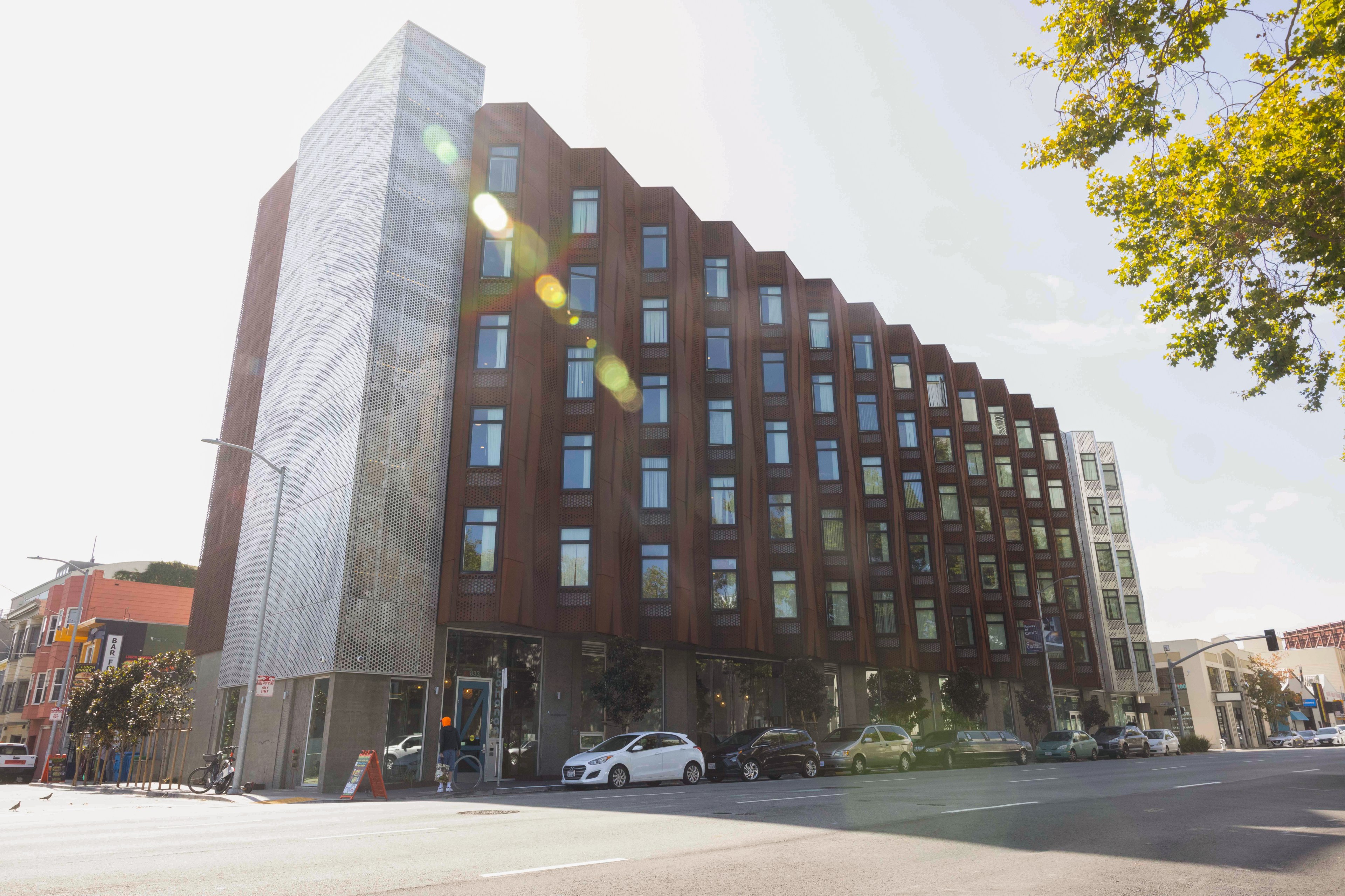 A modern, multi-story building with a distinct zigzag pattern on its brown facade and a perforated metal section on the left, sits along a tree-lined street.