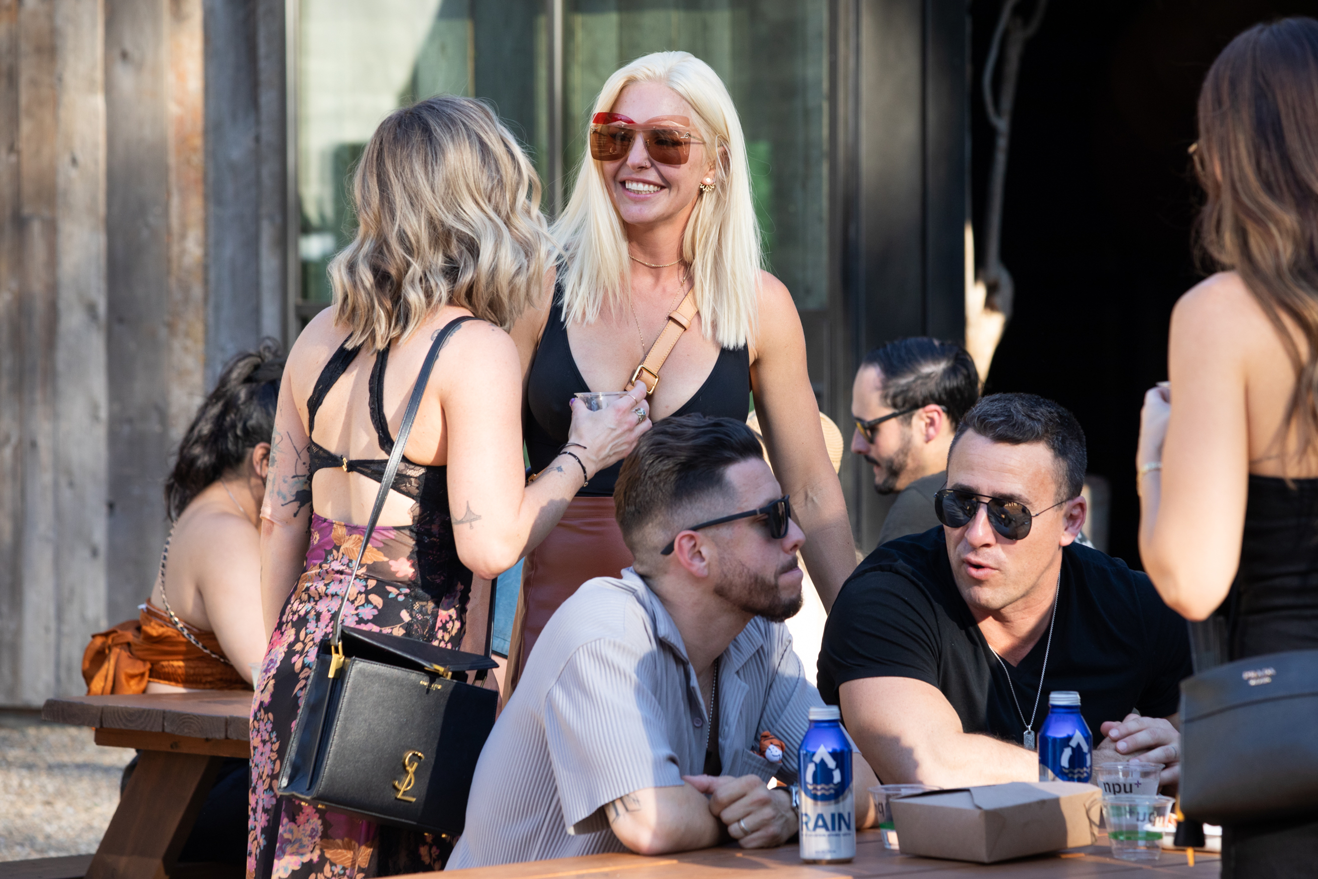 A group of friends is chatting outside, with two women standing and three men sitting at a wooden picnic table. They are casually dressed, wearing sunglasses, and enjoying beverages.