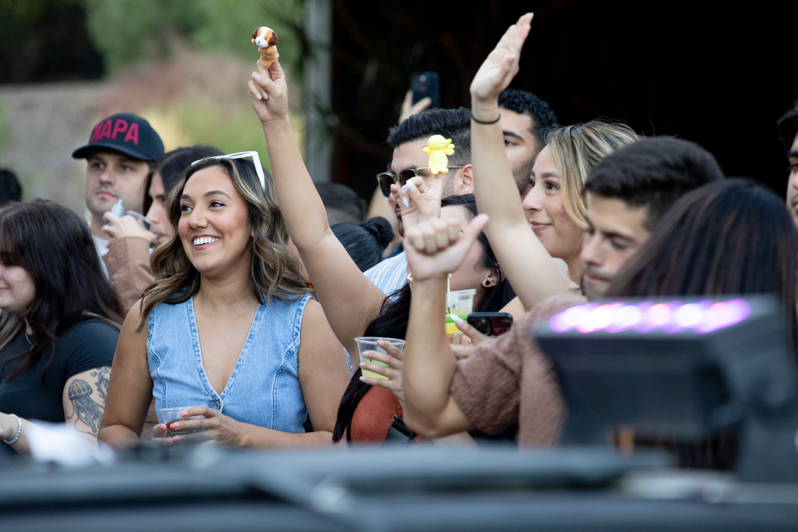 A smiling woman in a denim top is amid a crowd. People around her are raising hands and holding small toys, with one wearing a hat that says &quot;NAPA.&quot;