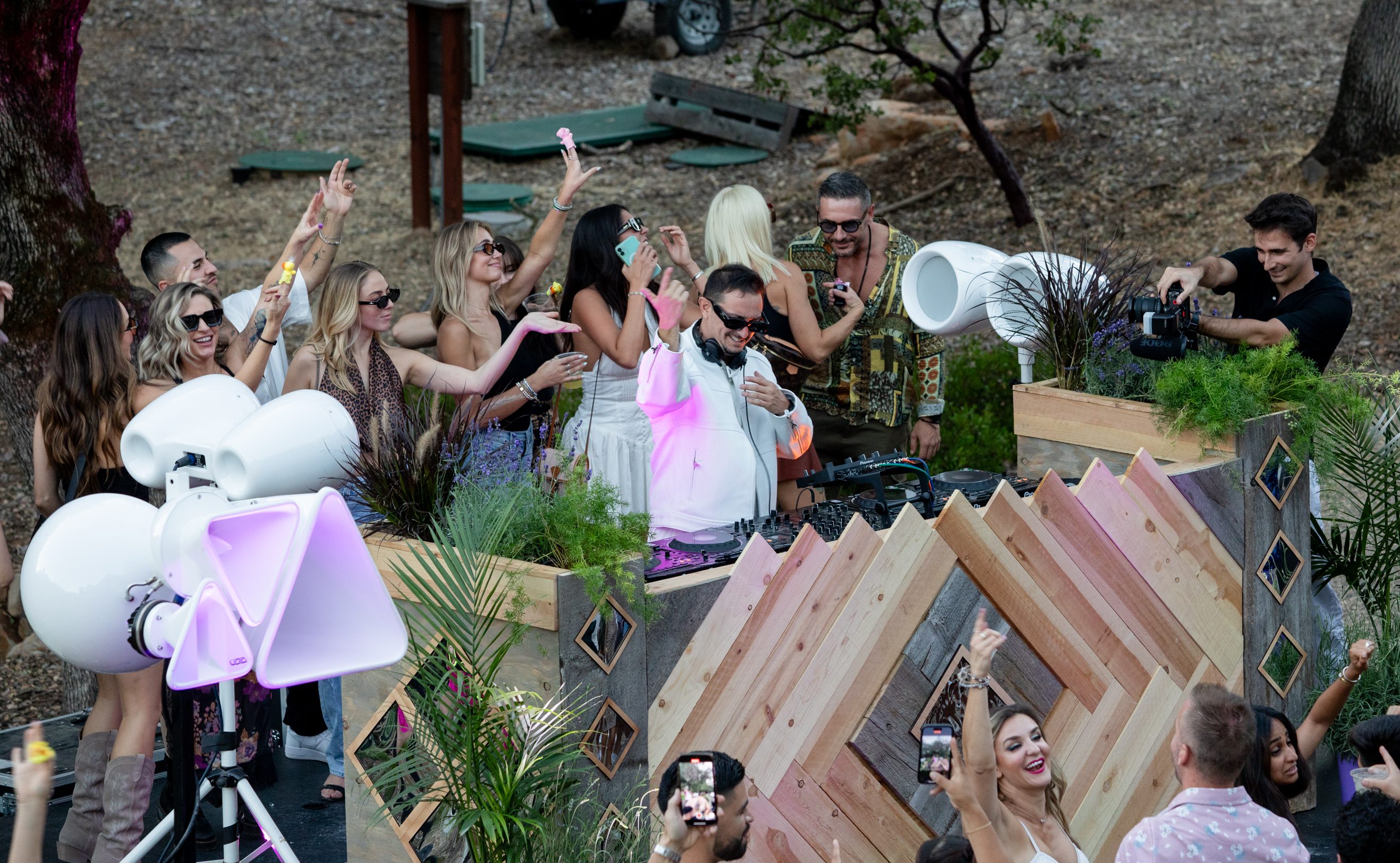 A lively outdoor music event with a DJ playing, surrounded by dancing and cheering people, modern decor, greenery, and a cameraman filming the crowd's excitement.