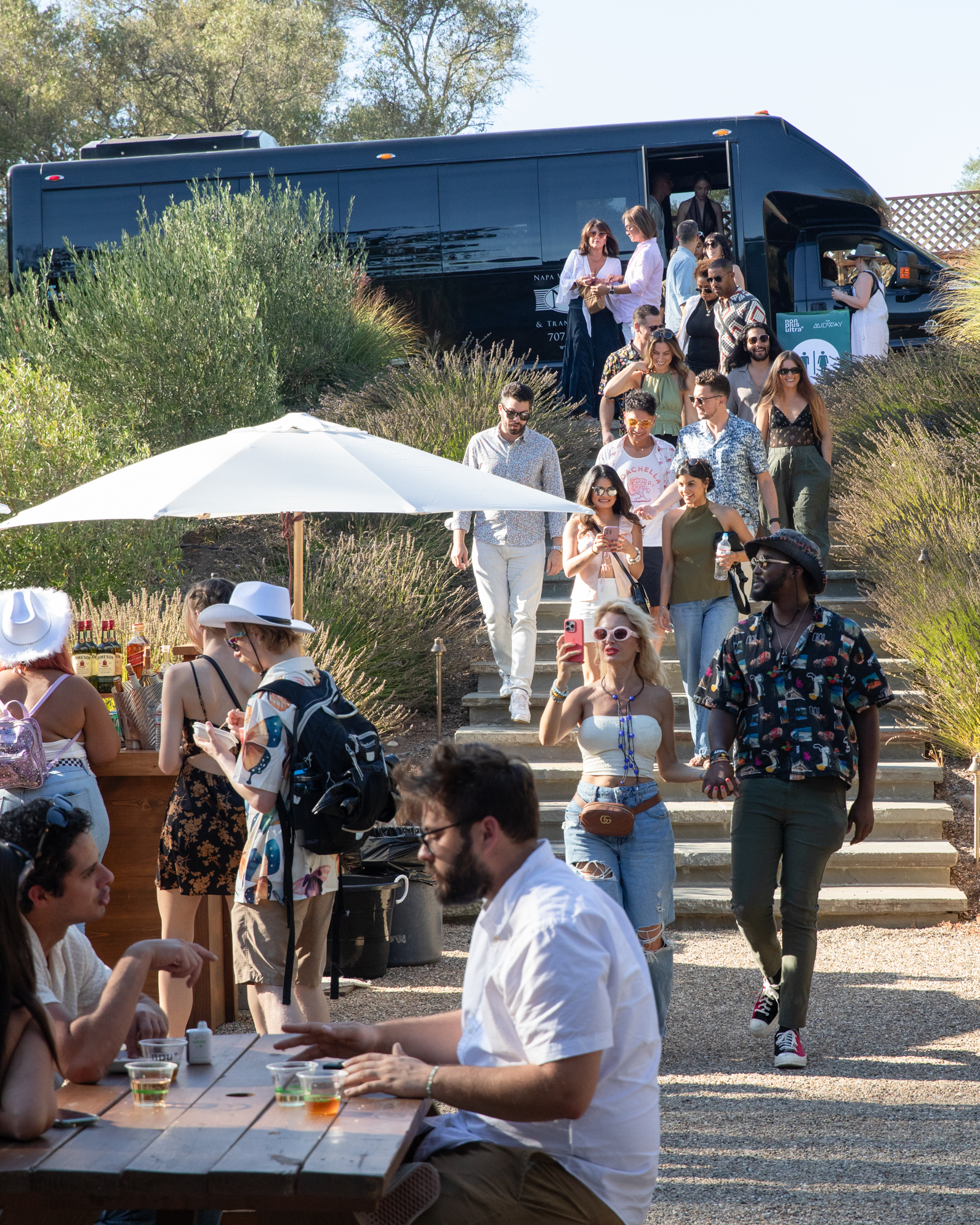 People are walking down steps from a large black bus in a garden setting. Others are sitting and standing near tables, some under a white umbrella, enjoying drinks and socializing.