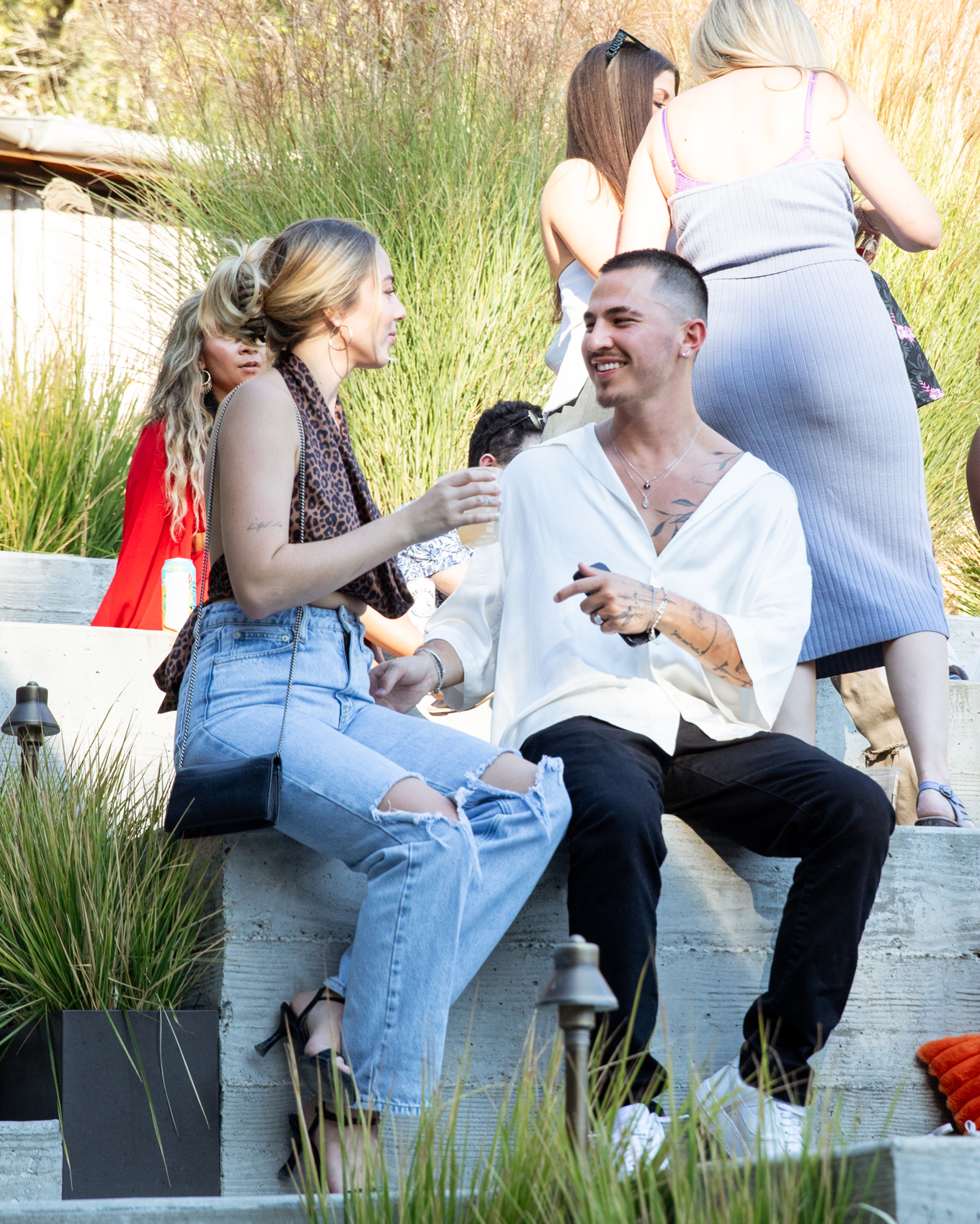 A smiling man and woman are seated on outdoor concrete steps, chatting. The scene includes other casually dressed people in the background, greenery, and soft sunlight.