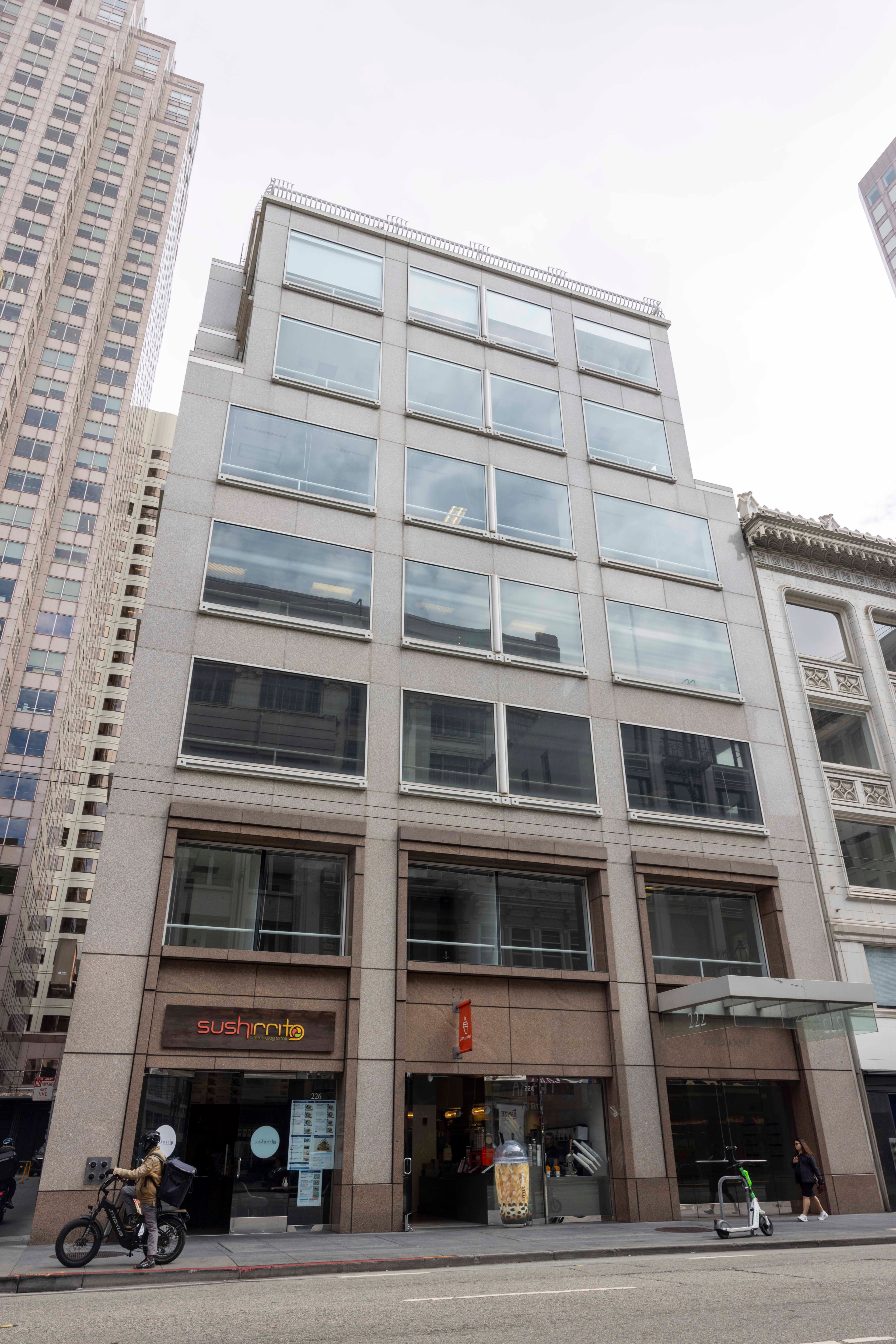 A modern, gray high-rise building with large windows houses a sushi restaurant and a bubble tea shop on the ground floor, located on a city street.