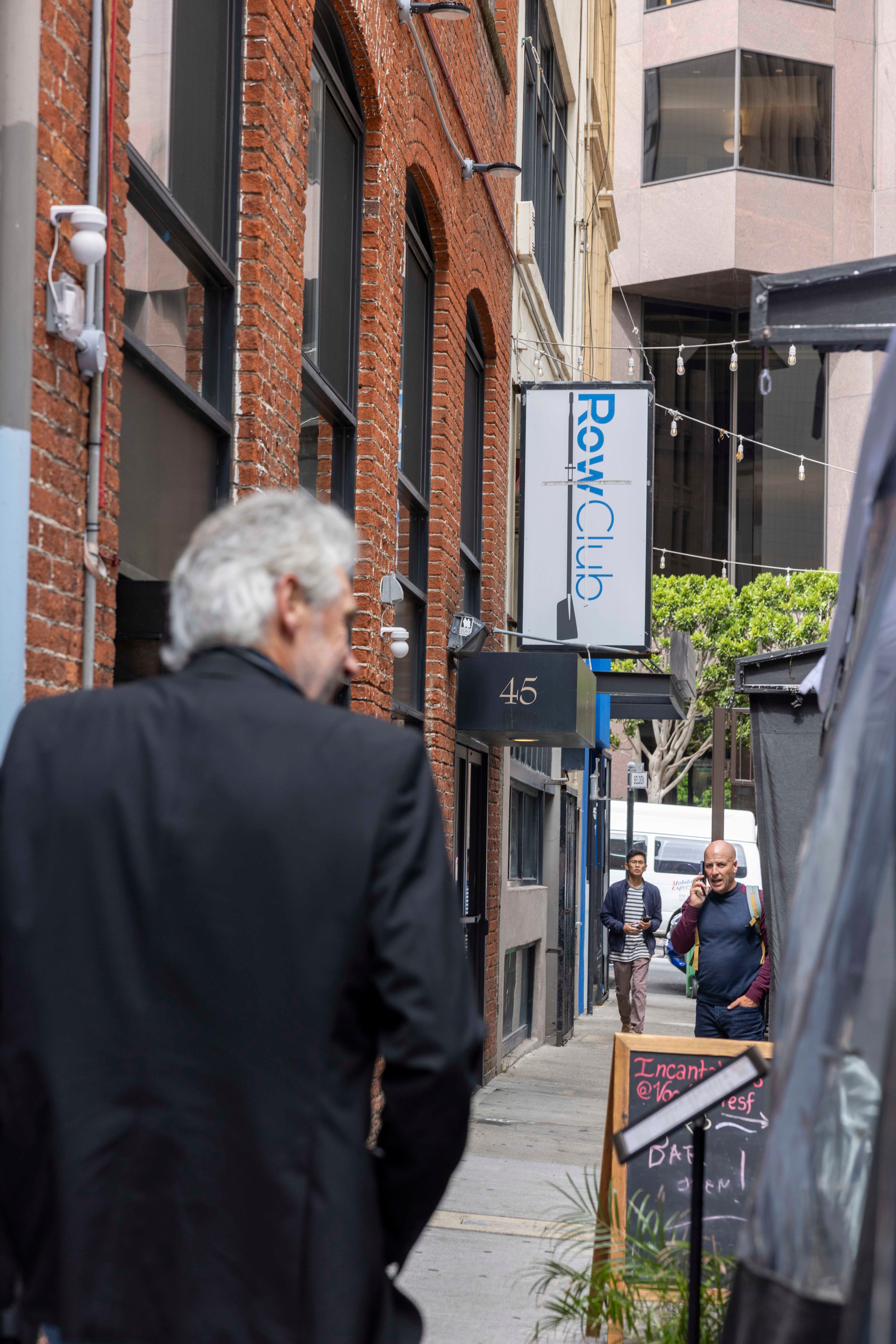 An alleyway with brick buildings features a sign for &quot;Row Club.&quot; Three men walk along the path, and a chalkboard sign stands on the right, partly obscured by plants.