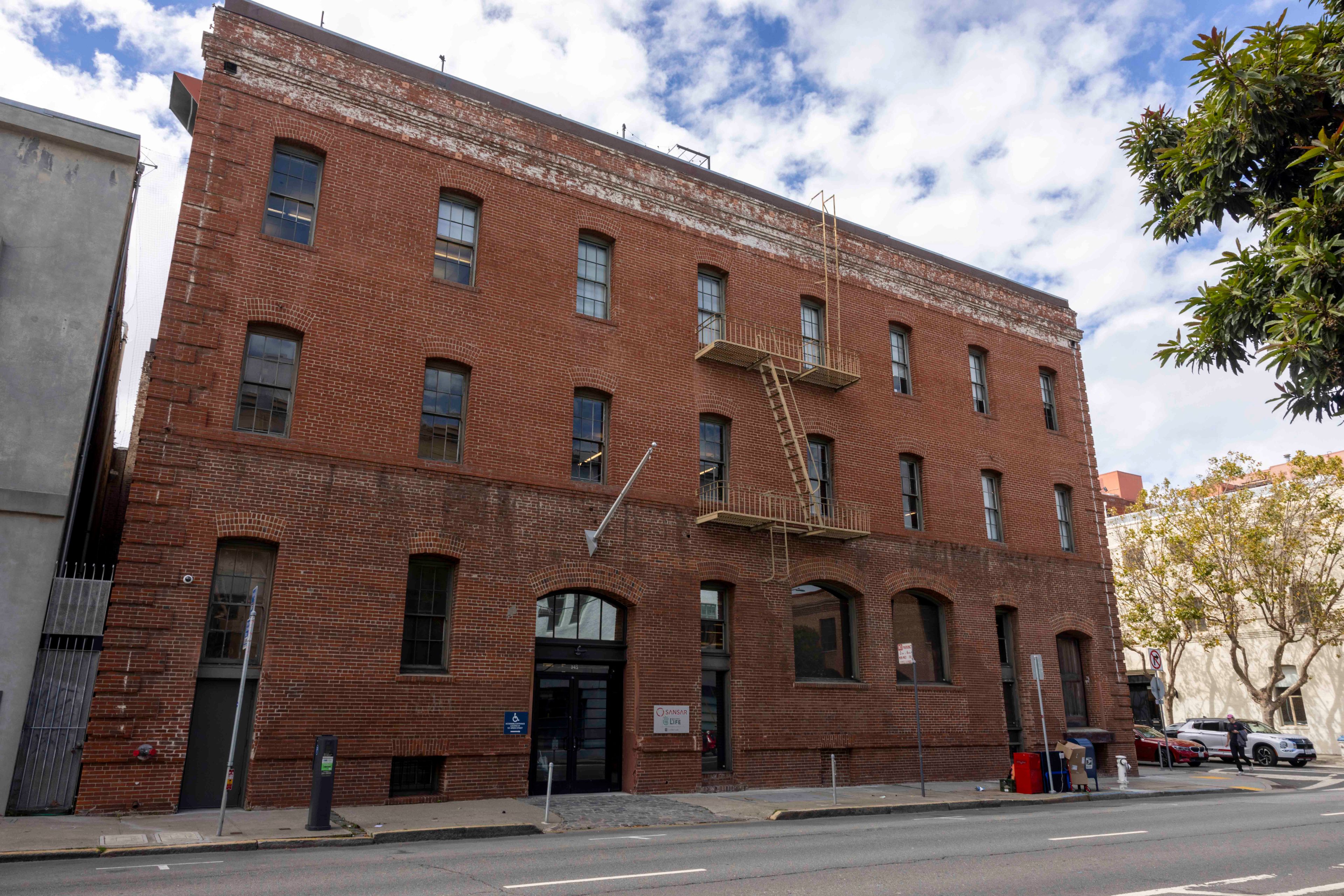 A multi-story brick building with tall windows, a fire escape, and a street in front. There's a tree on the right and another building nearby on the left.