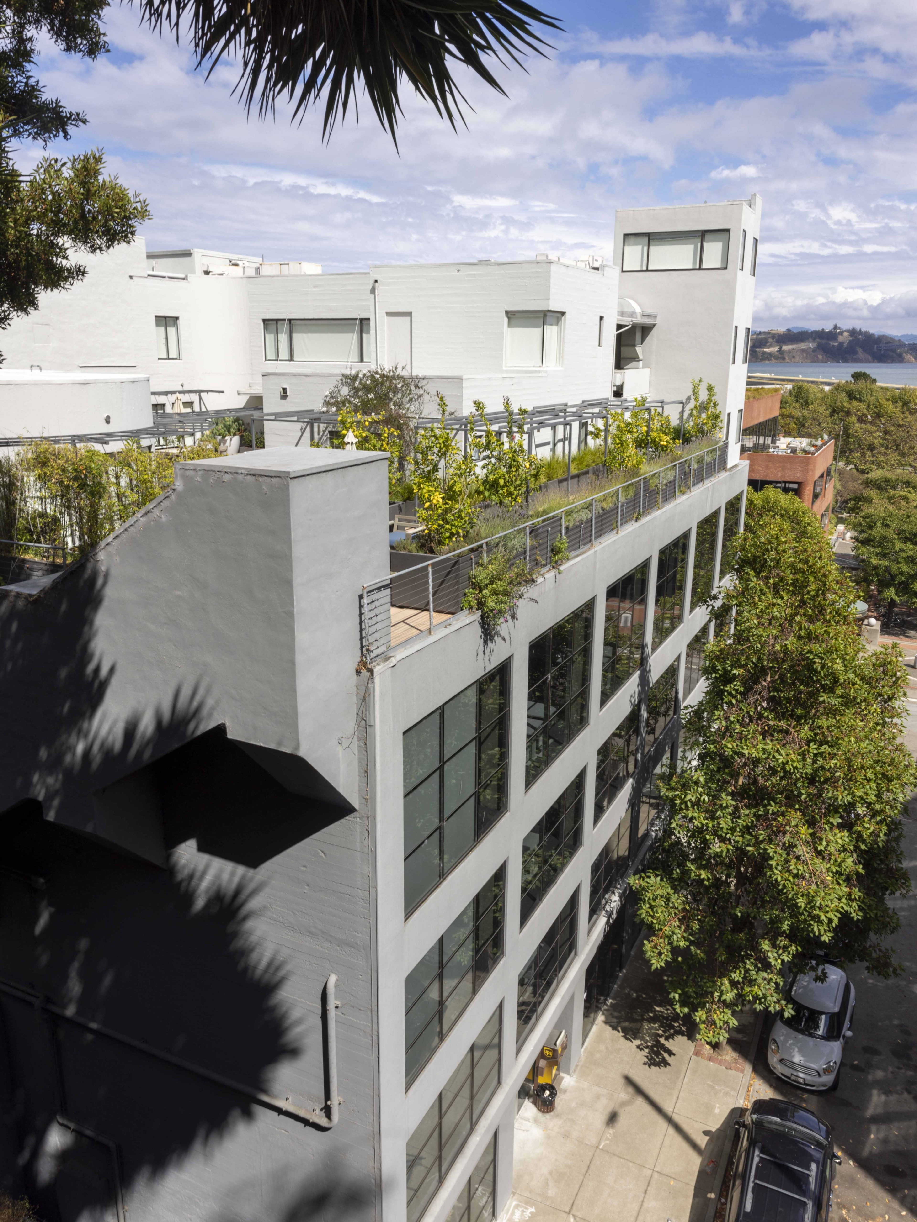 The image shows a modern building with large windows, an upper terrace with plants, and a backdrop of mountainous landscape and water under a partly cloudy sky.