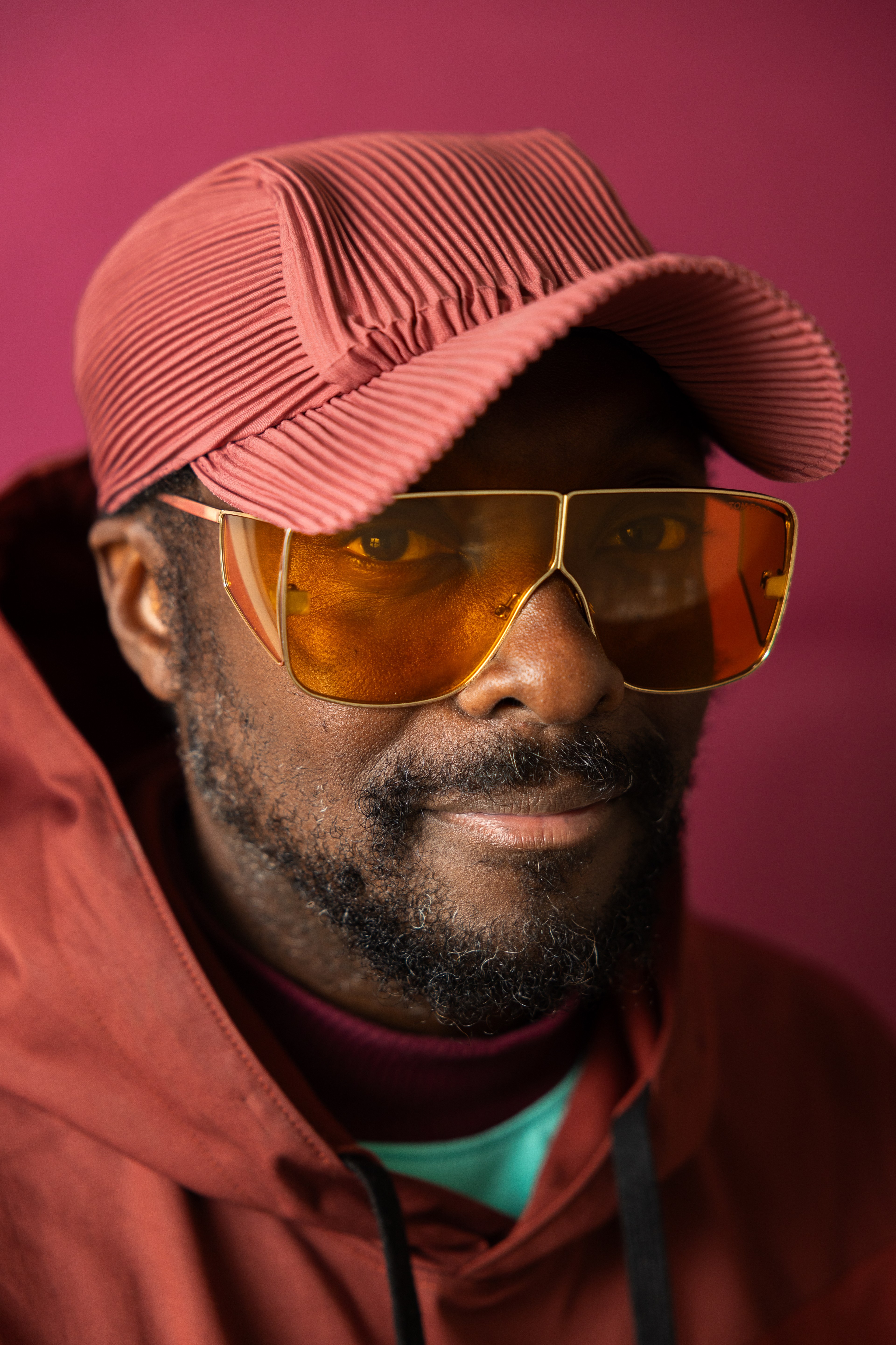 A man with a short beard wearing a textured pink cap, large gold-framed orange-tinted glasses, and a reddish jacket against a matching pink background.