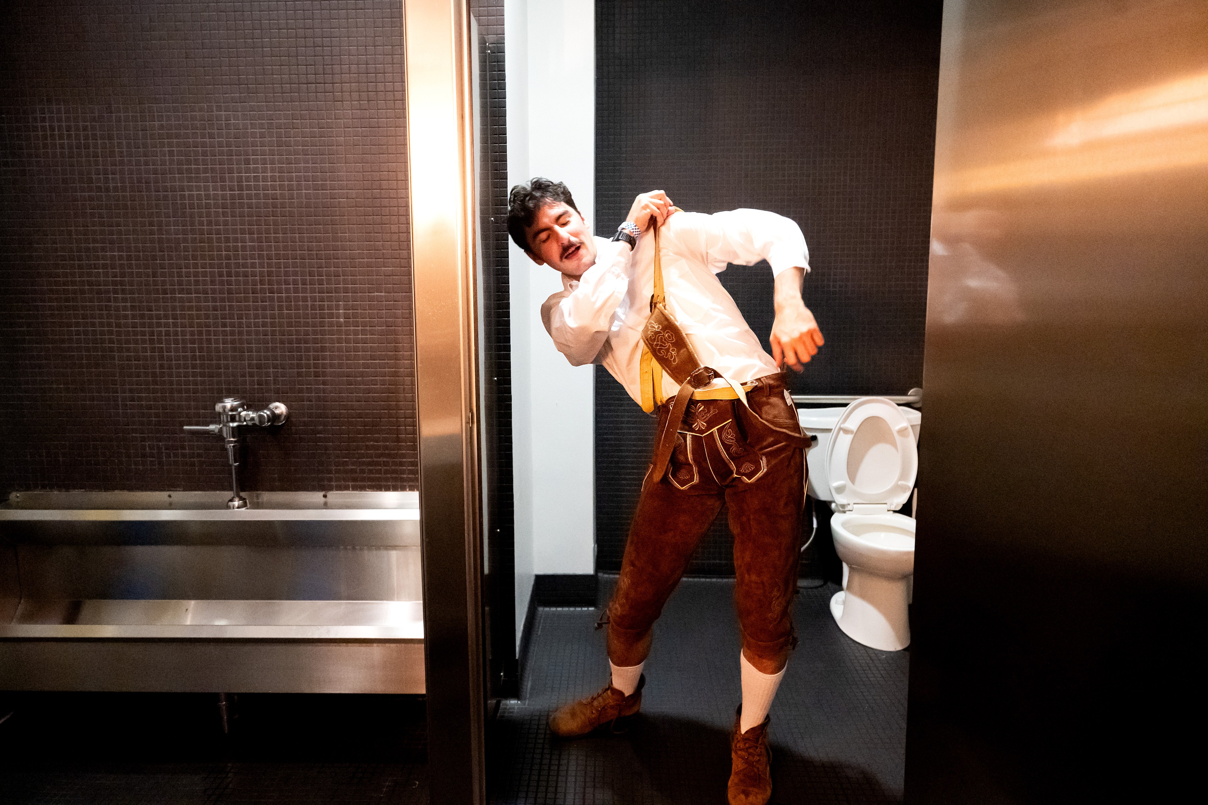 A man in Bavarian-style leather shorts and a white shirt poses comically in a bathroom with a urinal and toilet, against dark tiled walls.