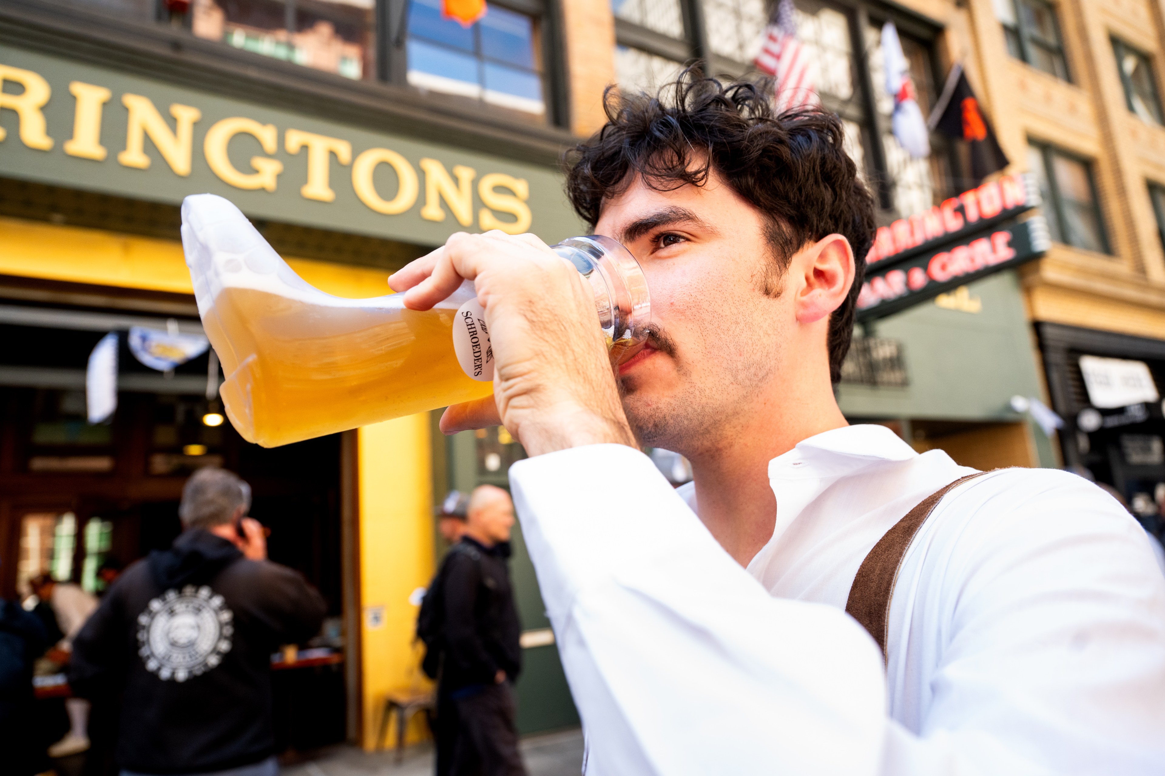 A man is drinking beer from a clear, boot-shaped glass outside a pub with "RINGTONS" and flags in the background, wearing a white shirt and brown suspenders.