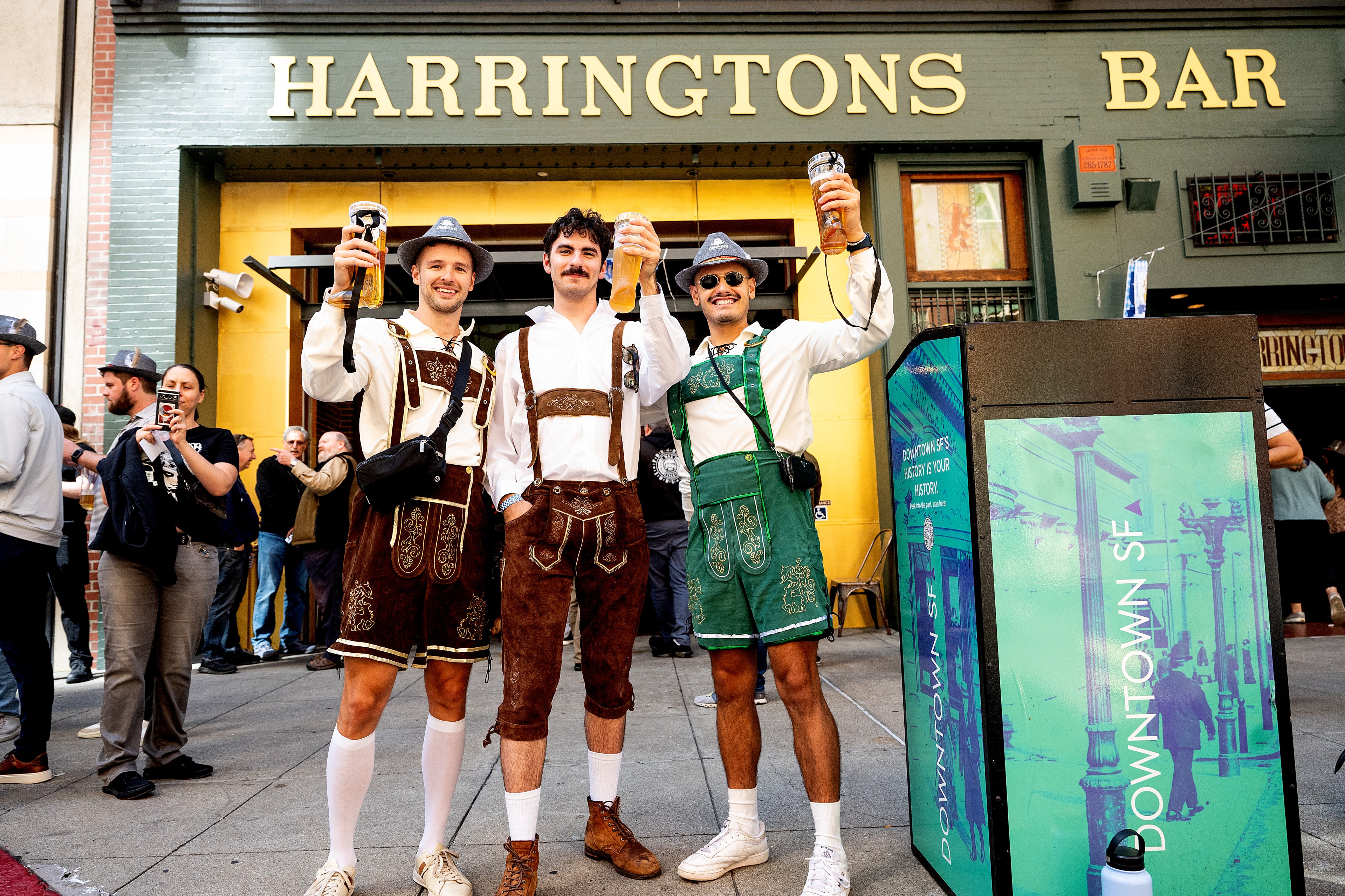 Three men dressed in traditional lederhosen hold up beers, smiling outside Harringtons Bar. The scene is lively with people in the background, capturing the festive atmosphere.