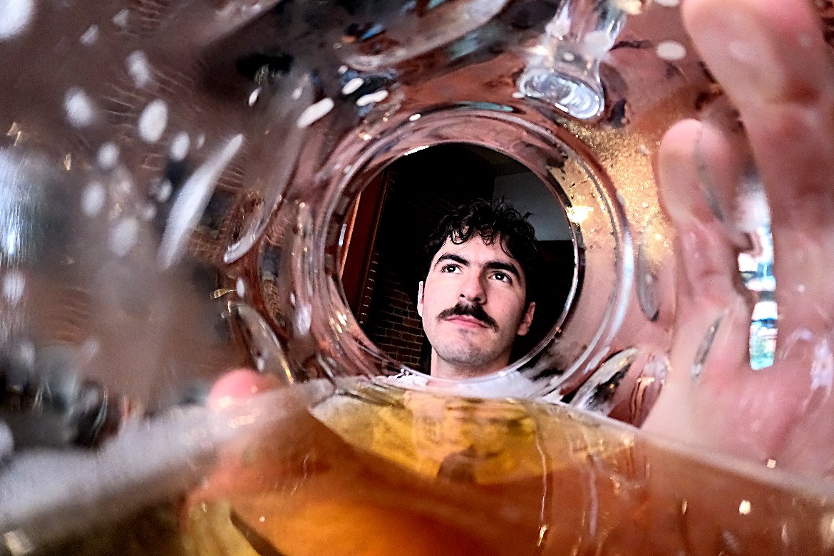 A man with a mustache is viewed through the bottom of a nearly empty beer glass, giving a distorted effect. His facial expression is thoughtful.