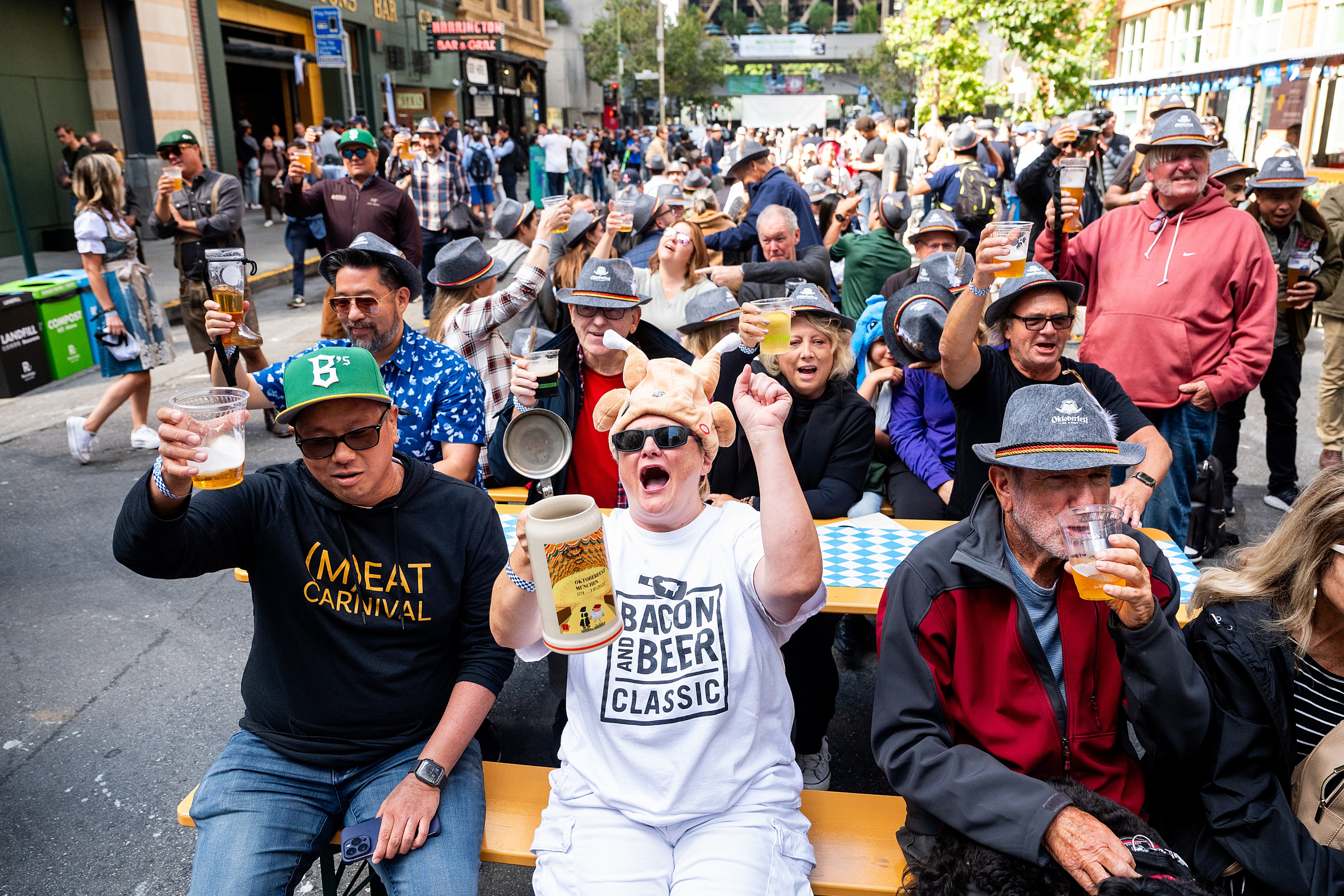 A lively crowd sits at long tables outdoors, holding mugs and cups of beer, and cheering. They wear festive attire, including hats and themed shirts.