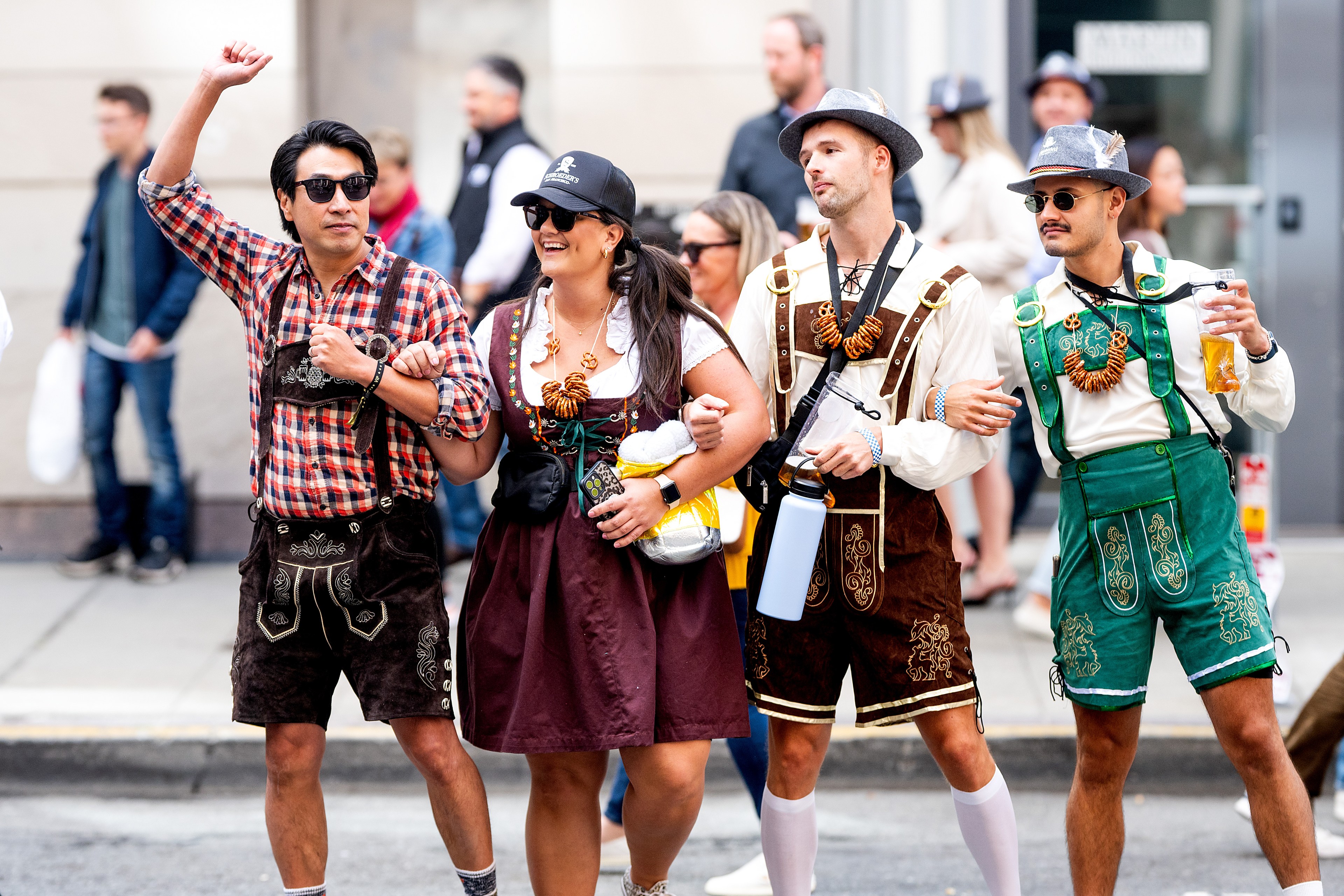 Four people in traditional Bavarian attire, including lederhosen and dirndls, walk arm-in-arm down a city sidewalk, smiling and enjoying a festive atmosphere.