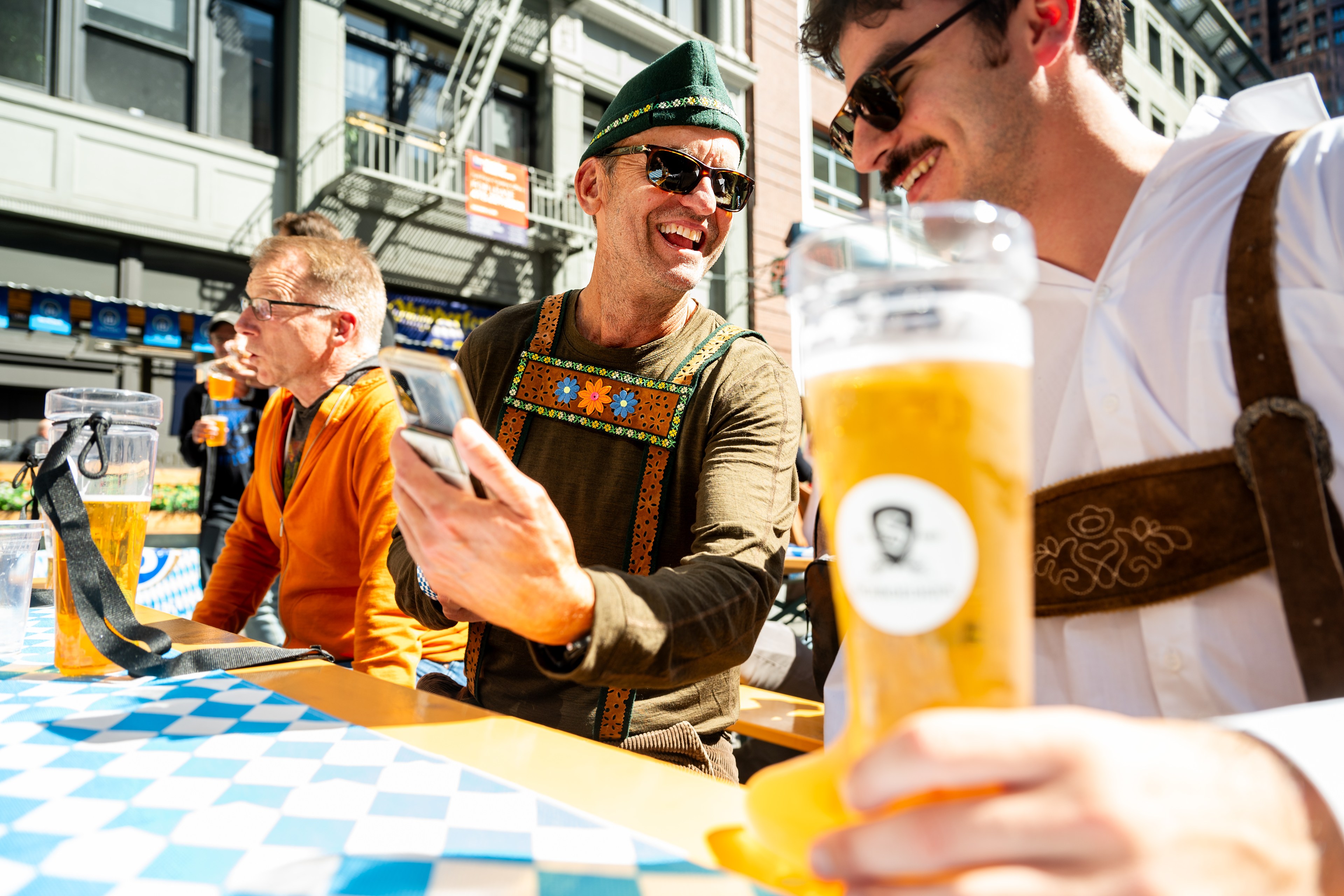 Three men dressed in traditional German attire are sitting outdoors, enjoying large glasses of beer, smiling, and engaged in conversation.