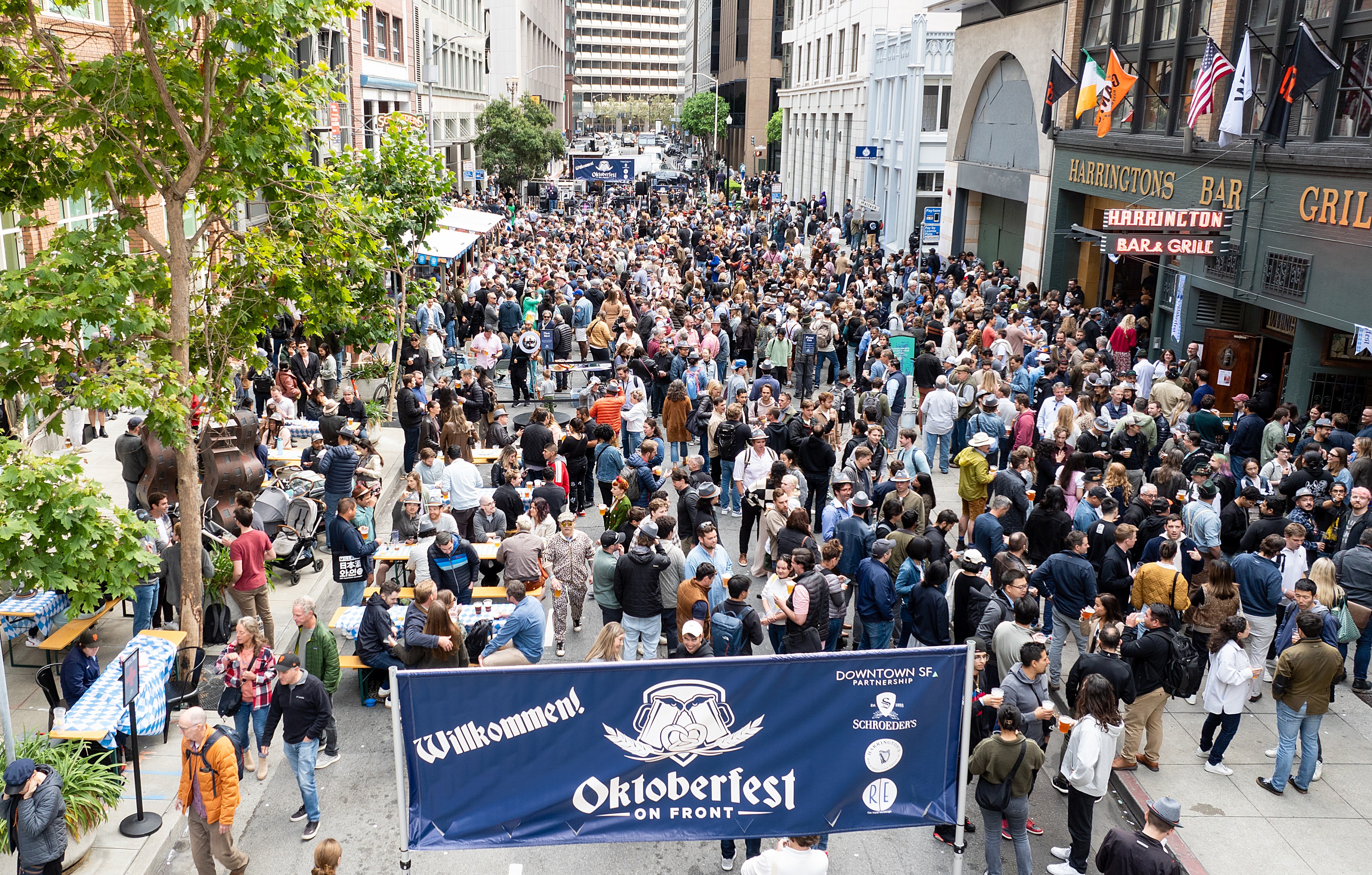 A bustling street filled with people at an outdoor Oktoberfest event, featuring tables, beer, and food stalls, while a large banner welcomes attendees.