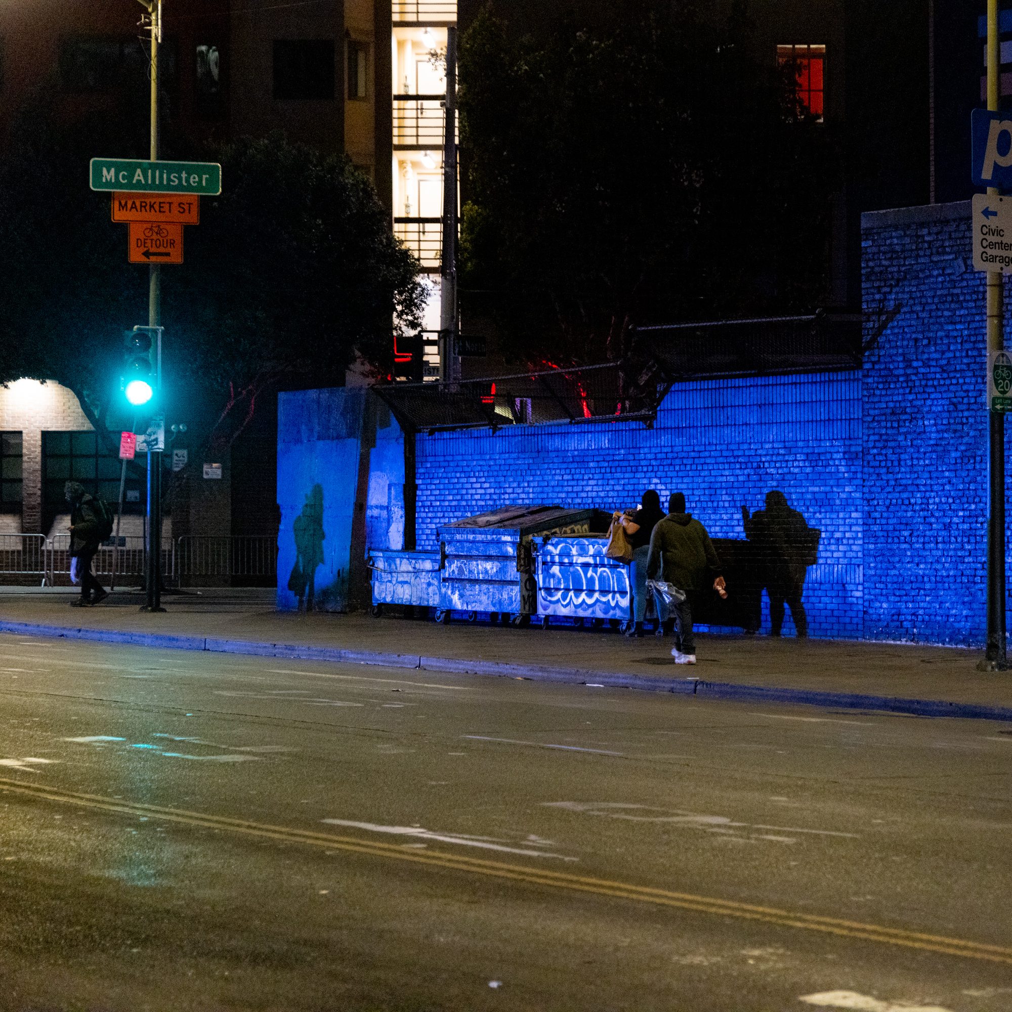 The image shows a nighttime urban scene with a lit street, a green traffic light, and signs for McAllister and Market St. People walk past a blue-lit wall and graffiti-covered dumpsters.