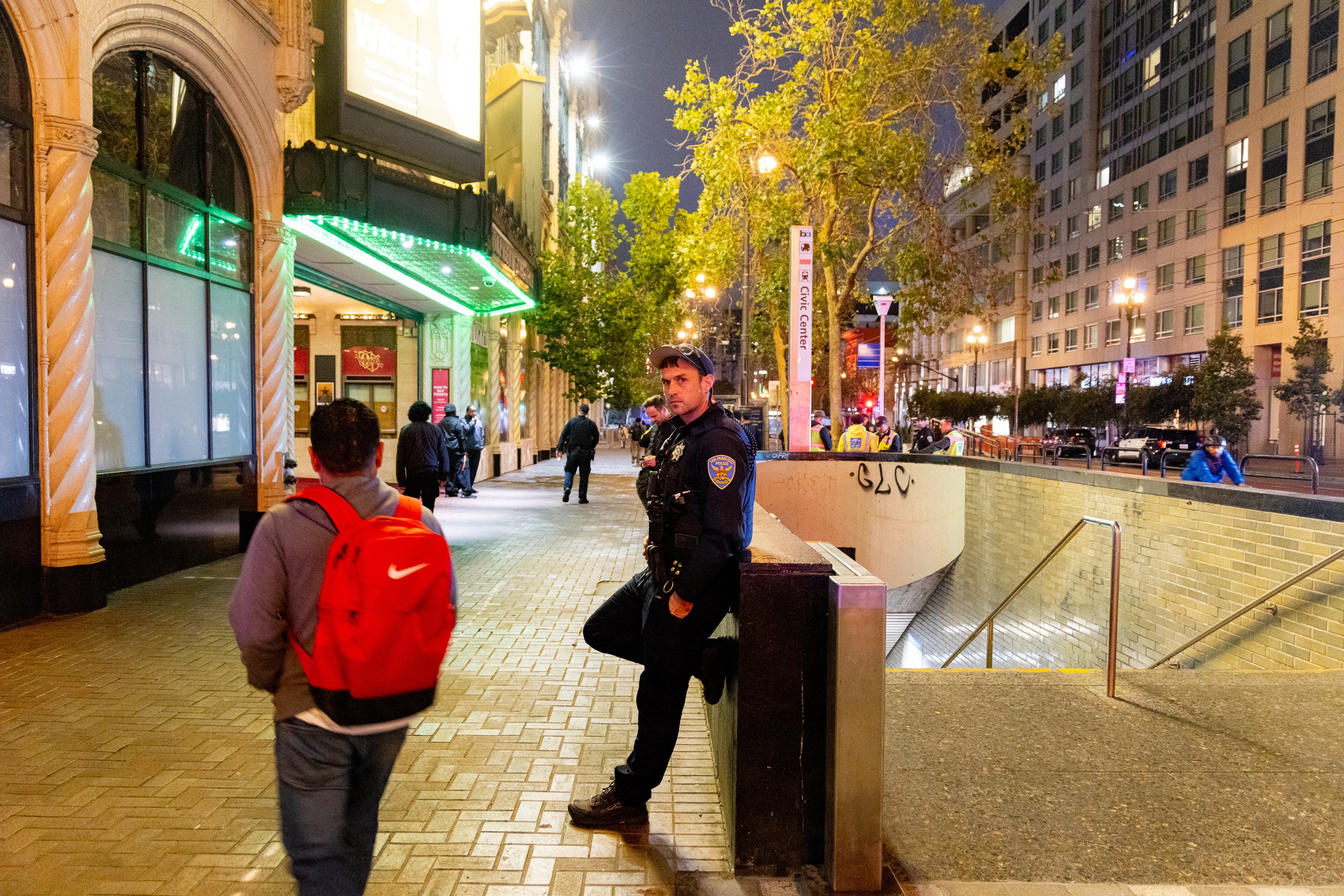 A police officer stands beside a subway entrance in a city at night, while pedestrians walk along the sidewalk near a brightly lit building with green lights.