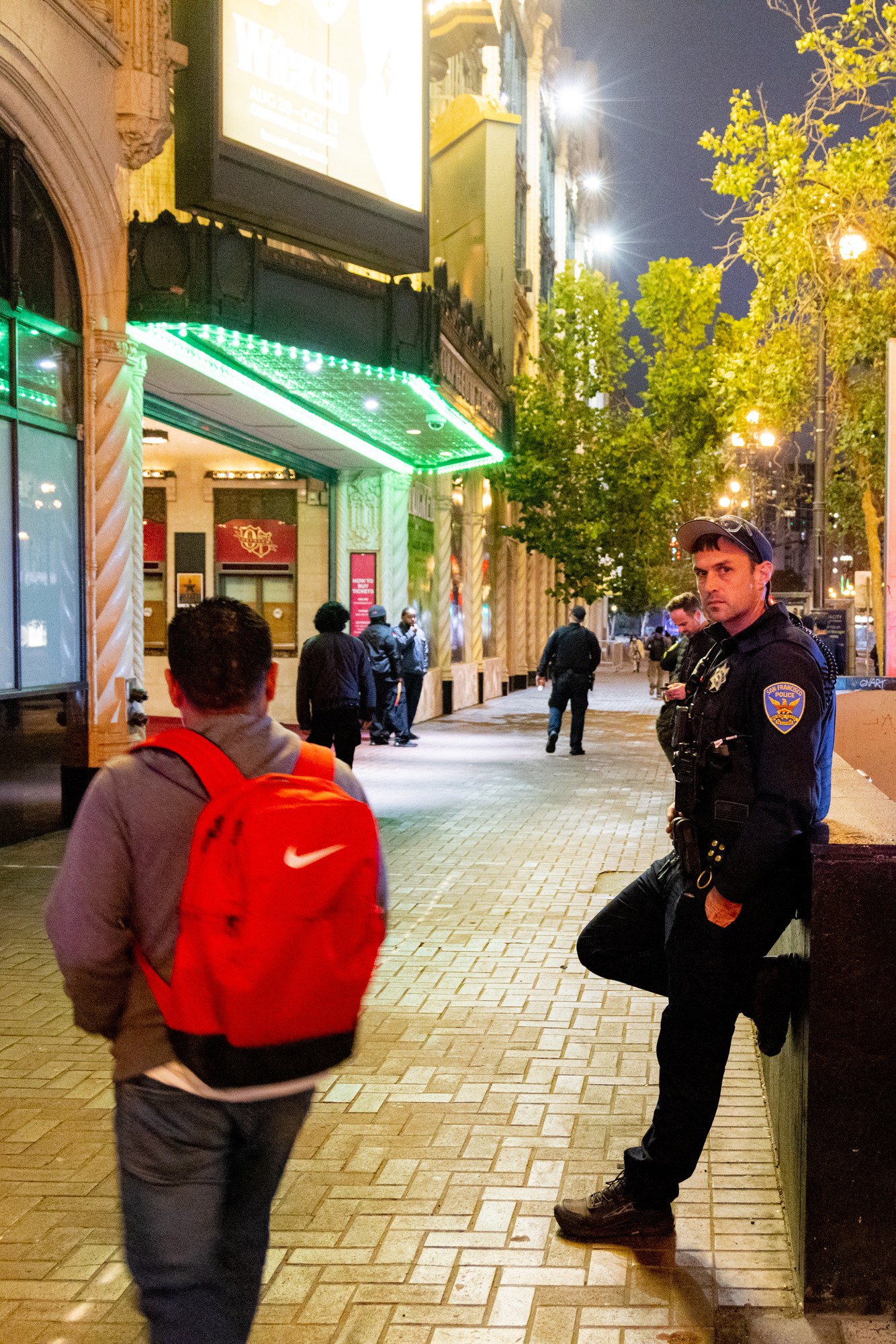 A police officer stands beside a subway entrance in a city at night, while pedestrians walk along the sidewalk near a brightly lit building with green lights.