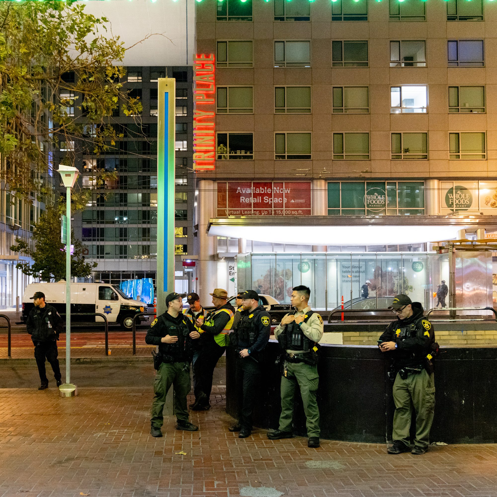 A group of law enforcement officers are gathered near a building with &quot;TRINITY PLACE&quot; sign. They seem to be patrolling or discussing, with one officer standing apart.